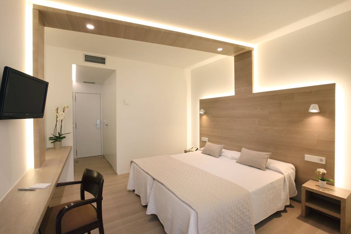 A modern hotel room with a large bed, a desk with a chair, a TV, and a small bedside table with a lamp.