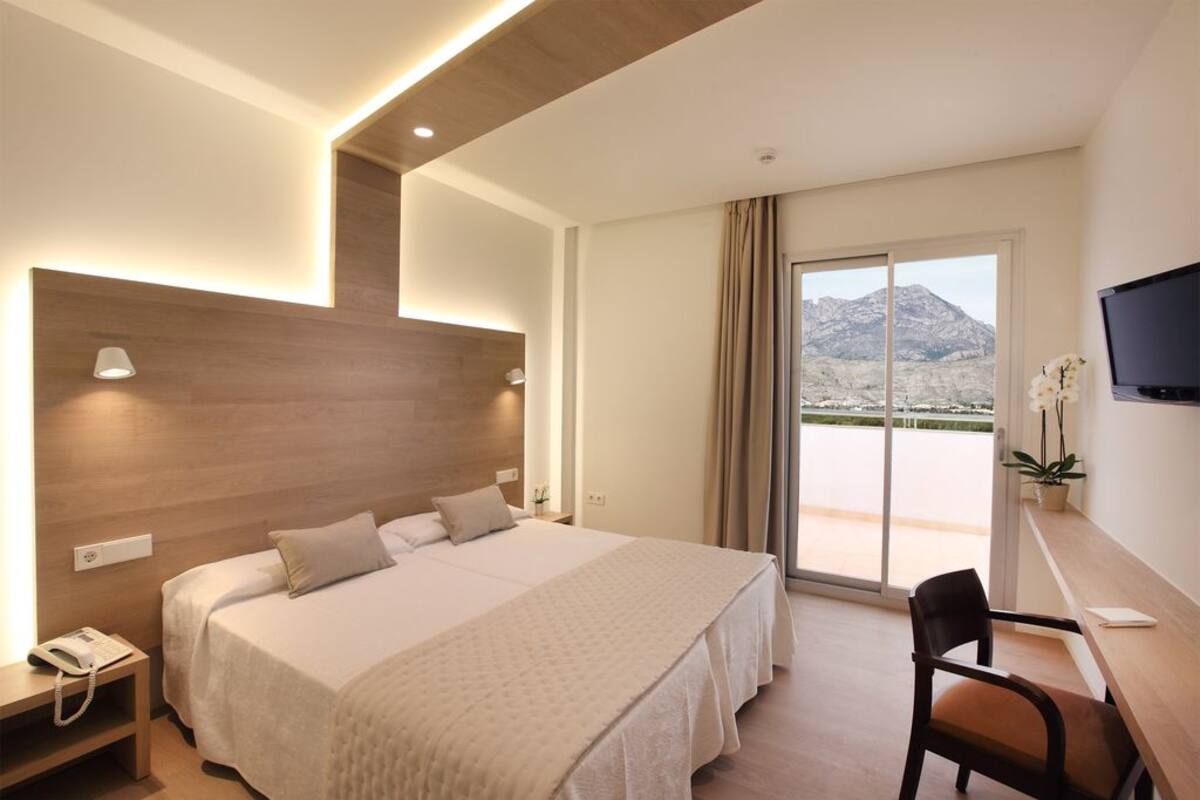 A hotel room with a large bed, a desk, a chair, a TV, and a view of a mountain.