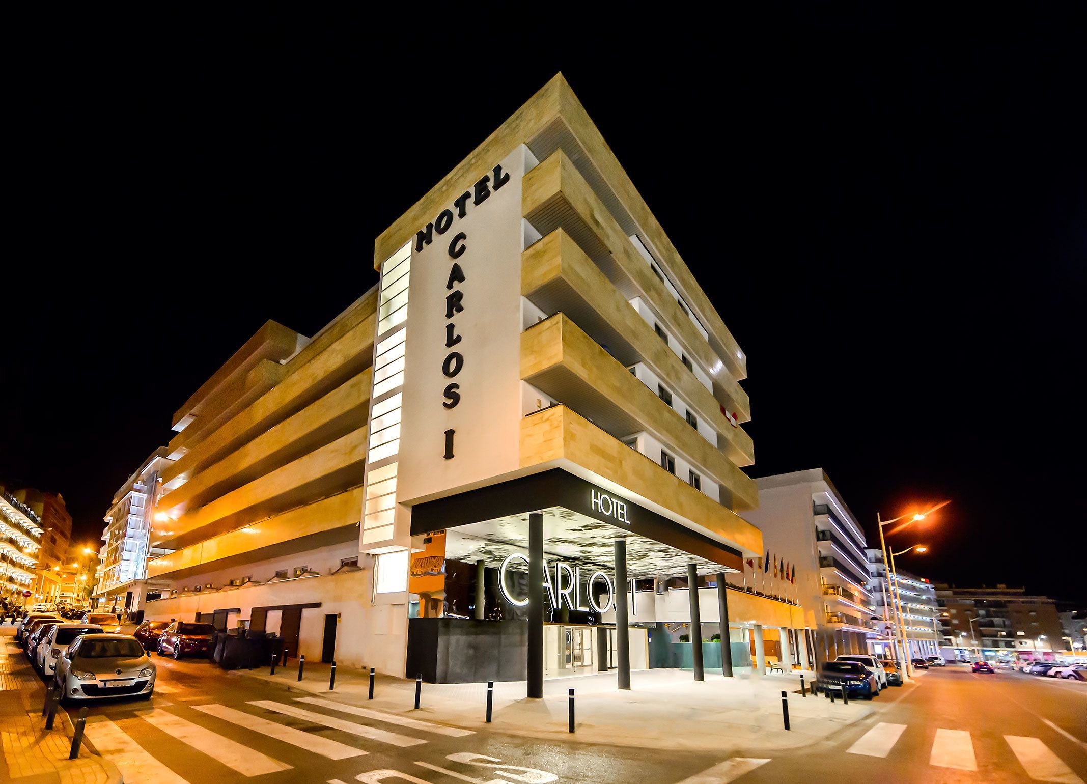 Een meerlaags hotel met een opvallend 'HOTEL CARLOS I' bord, met een modern architectonisch ontwerp, een plat dak en meerdere balkons.