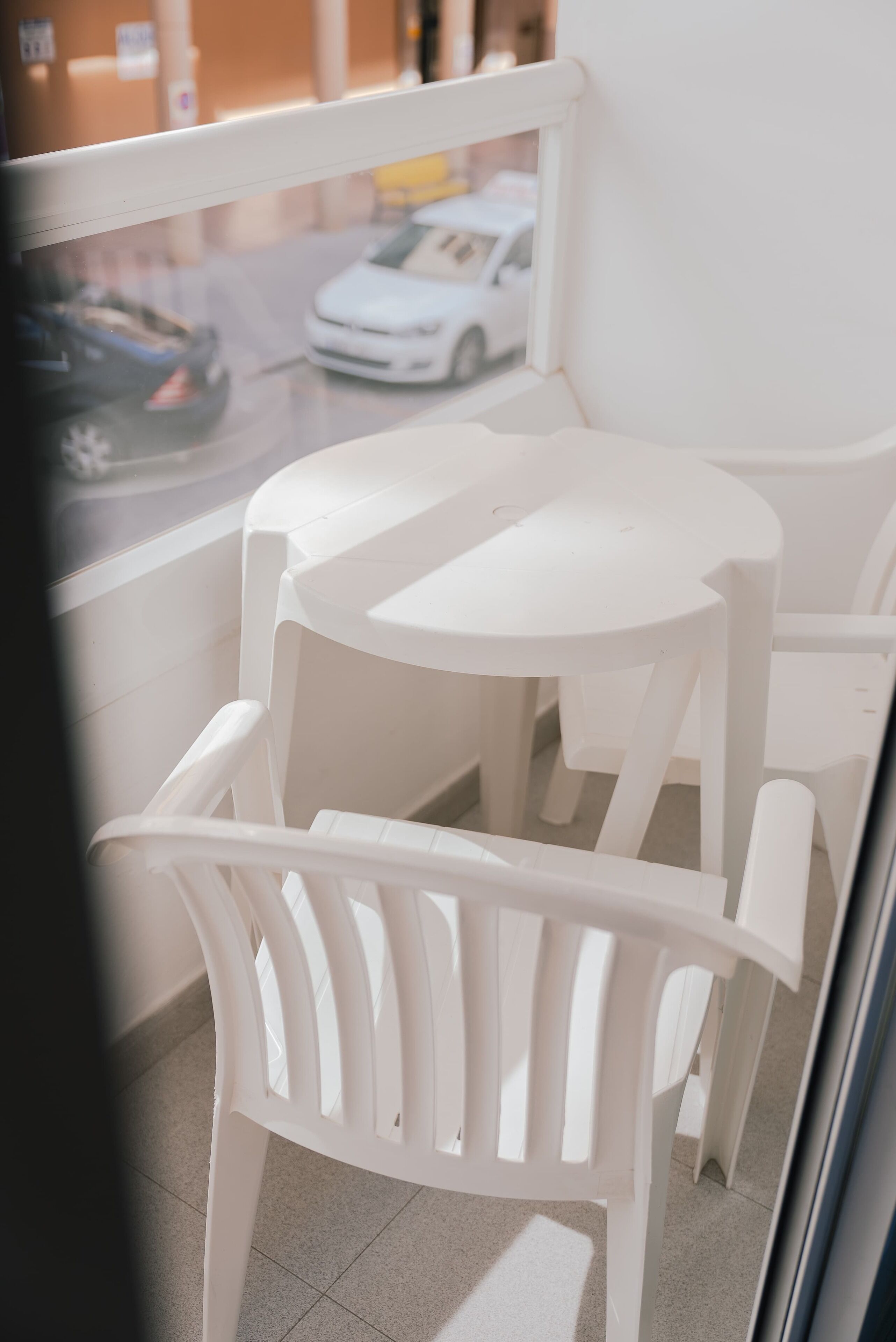 Un balcón con sillas y una mesa de plástico blanco, con vista a una calle donde hay coches aparcados.