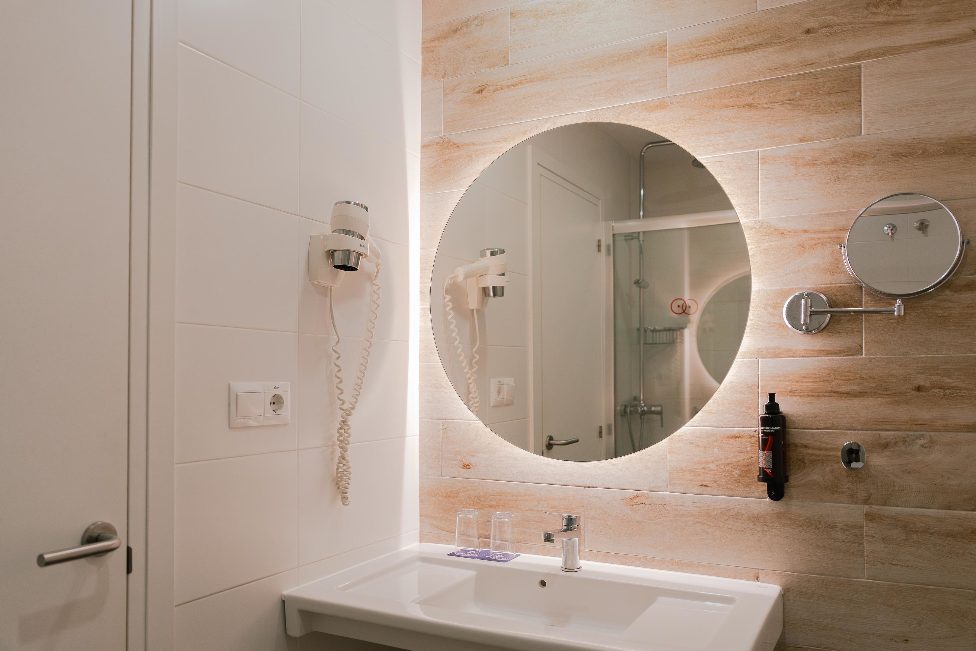 Un baño con un lavamanos blanco, un espejo redondo y un secador de pelo fijado en la pared.