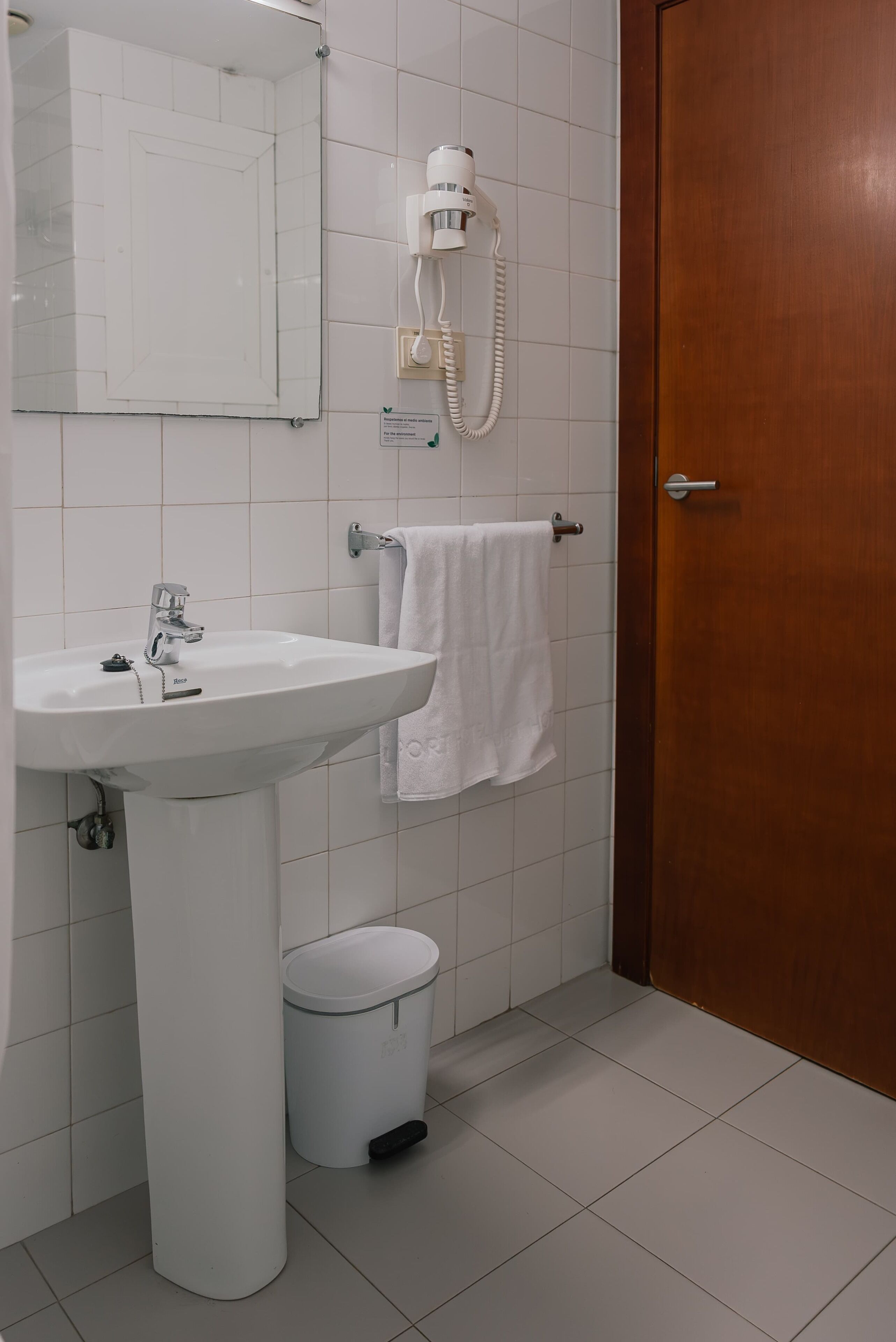 A bathroom with a white sink, a mirror, a hairdryer, a towel rack, and a wooden door.