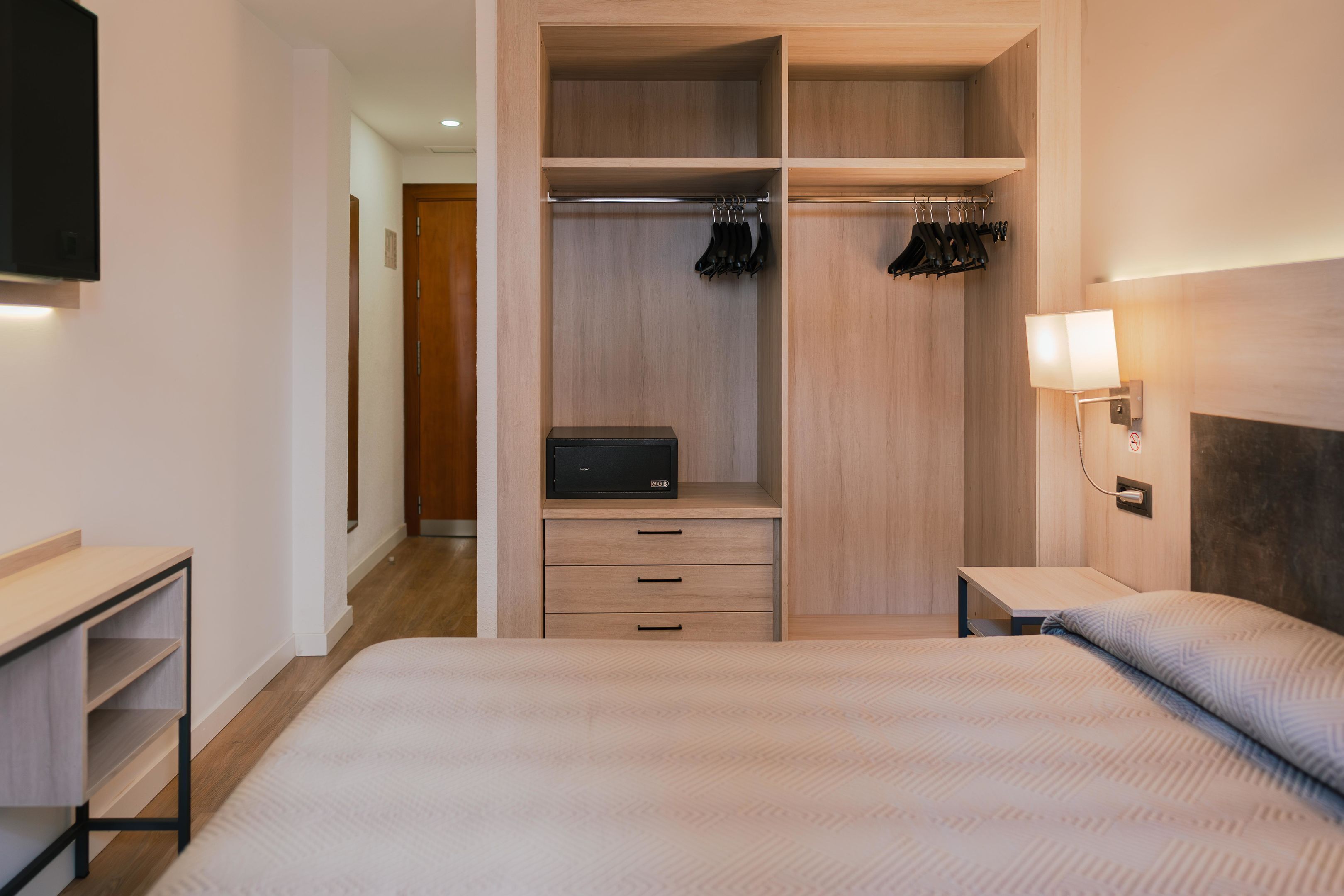 Habitación de hotel con cama, armario de madera con espacio para colgar, mesita de noche y lámpara de pared.