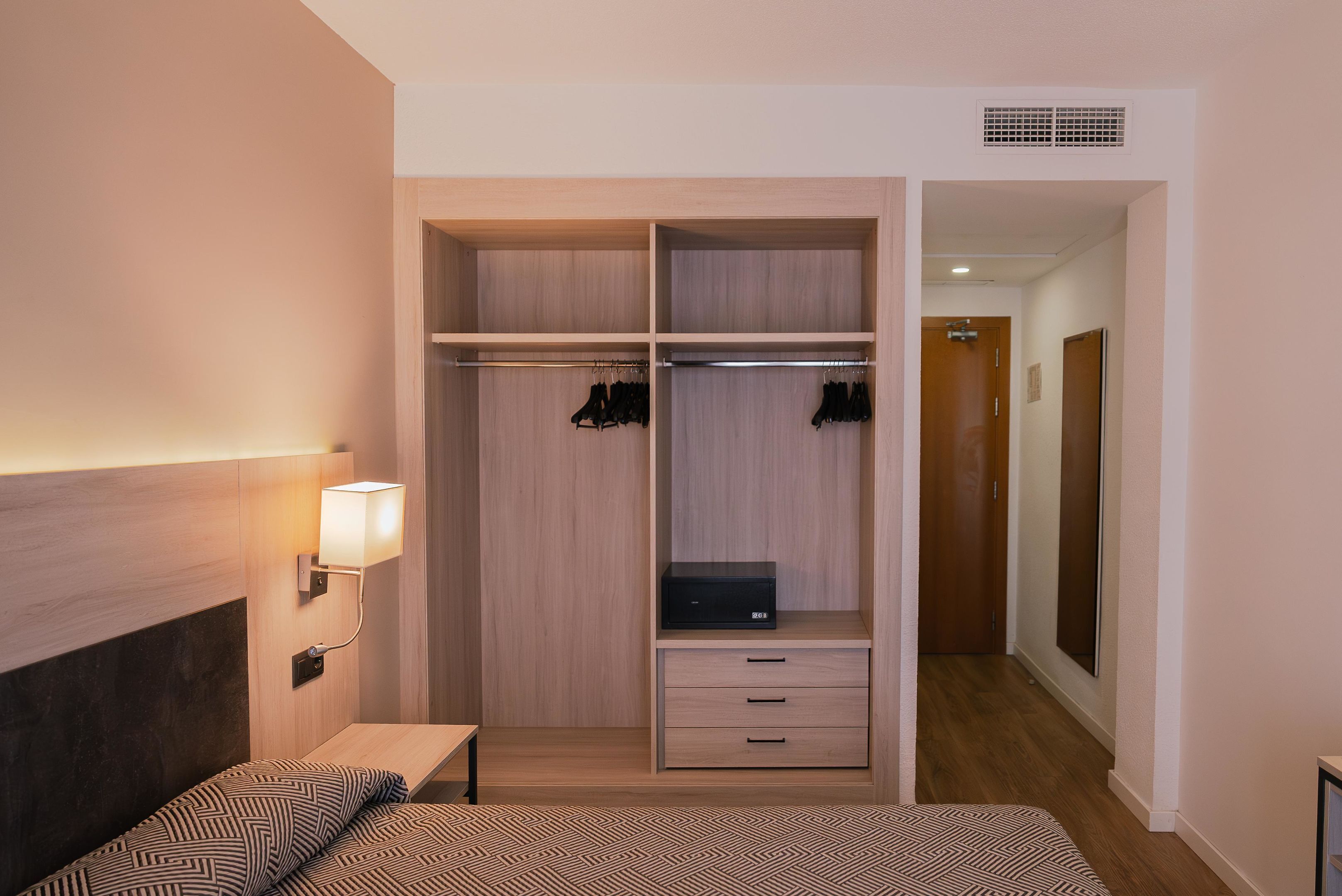 Une chambre d’hôtel comprenant un lit, une table de chevet, une armoire avec des vêtements suspendus, un bureau avec des tiroirs et une lampe fixée au mur.