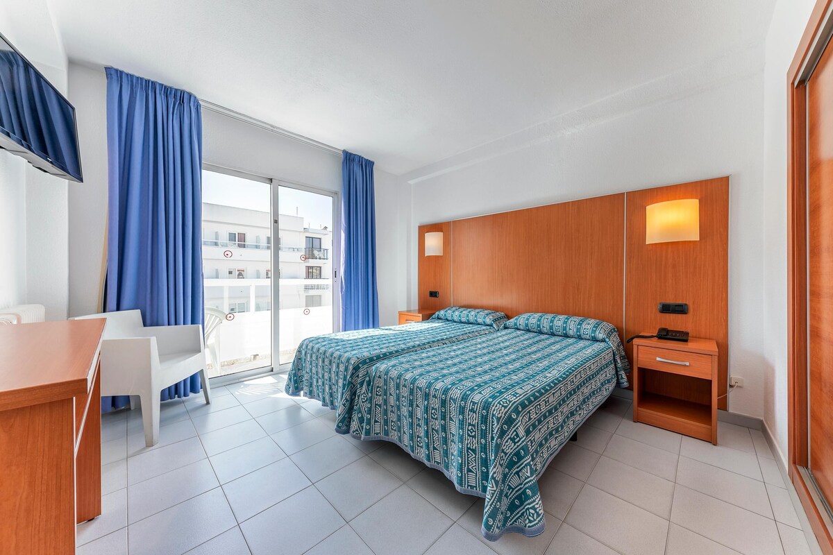 A modern hotel room with a large bed, a desk, and a chair. There is a balcony with blue curtains.