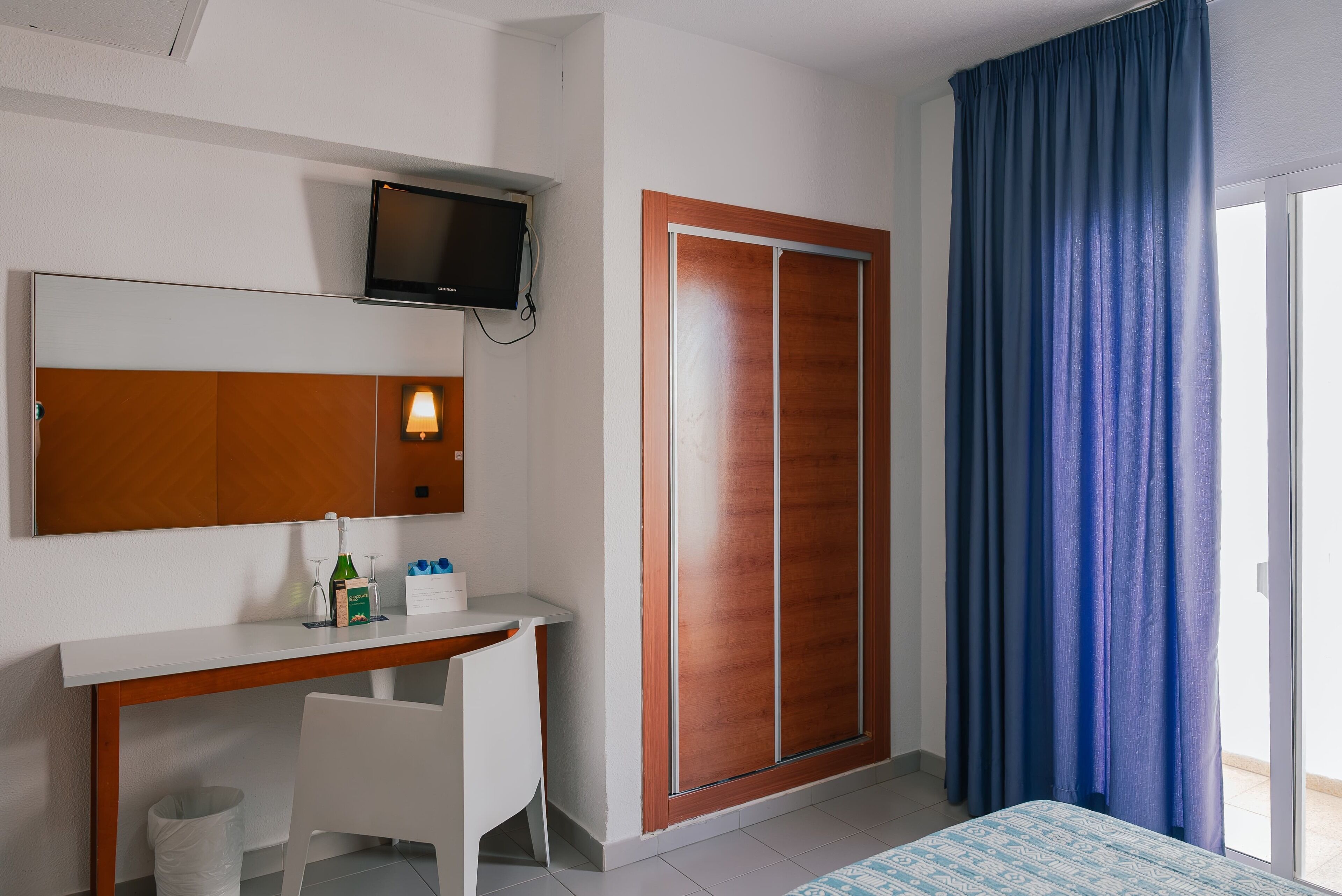 A hotel room with a wooden desk, a television mounted on the wall, a wardrobe, and a large window with blue curtains.