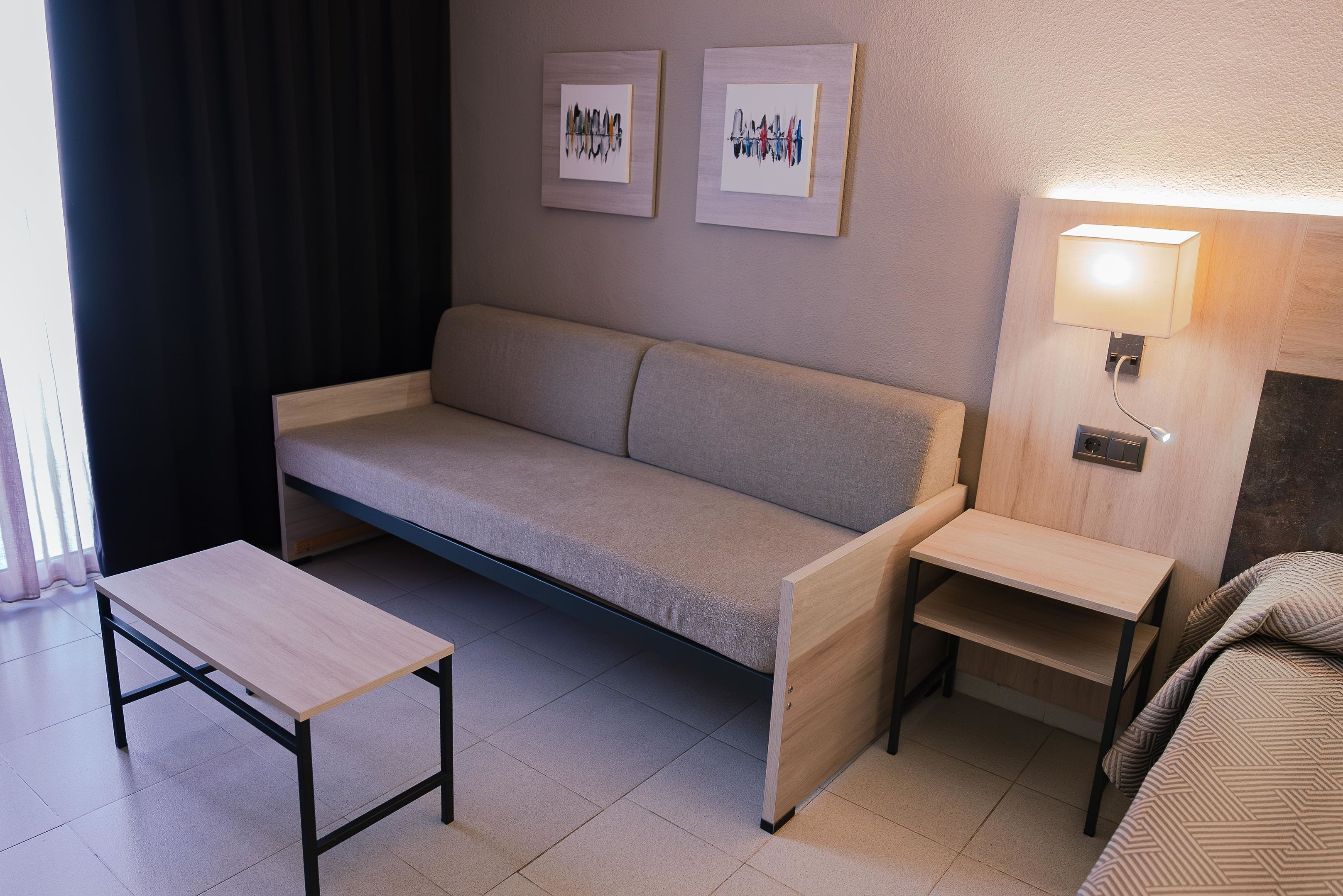 Une chambre d’hôtel compacte comprenant un canapé, une petite table, une table de chevet avec une lampe, et deux œuvres d’art encadrées accrochées au mur.