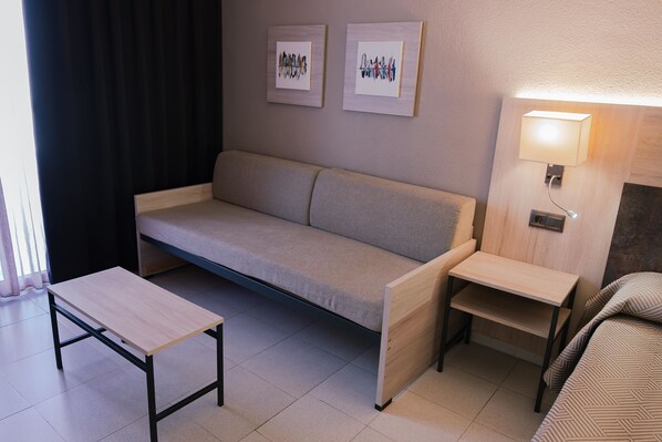 Habitación de hotel pequeña y funcional, con sofá, una mesita, una mesita de noche con lámpara y dos cuadros enmarcados en la pared.