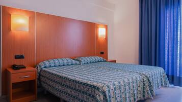 Una habitación de hotel con cabecera de madera, cubrecama estampado, mesita de noche con teléfono y ventana con cortinas azules.