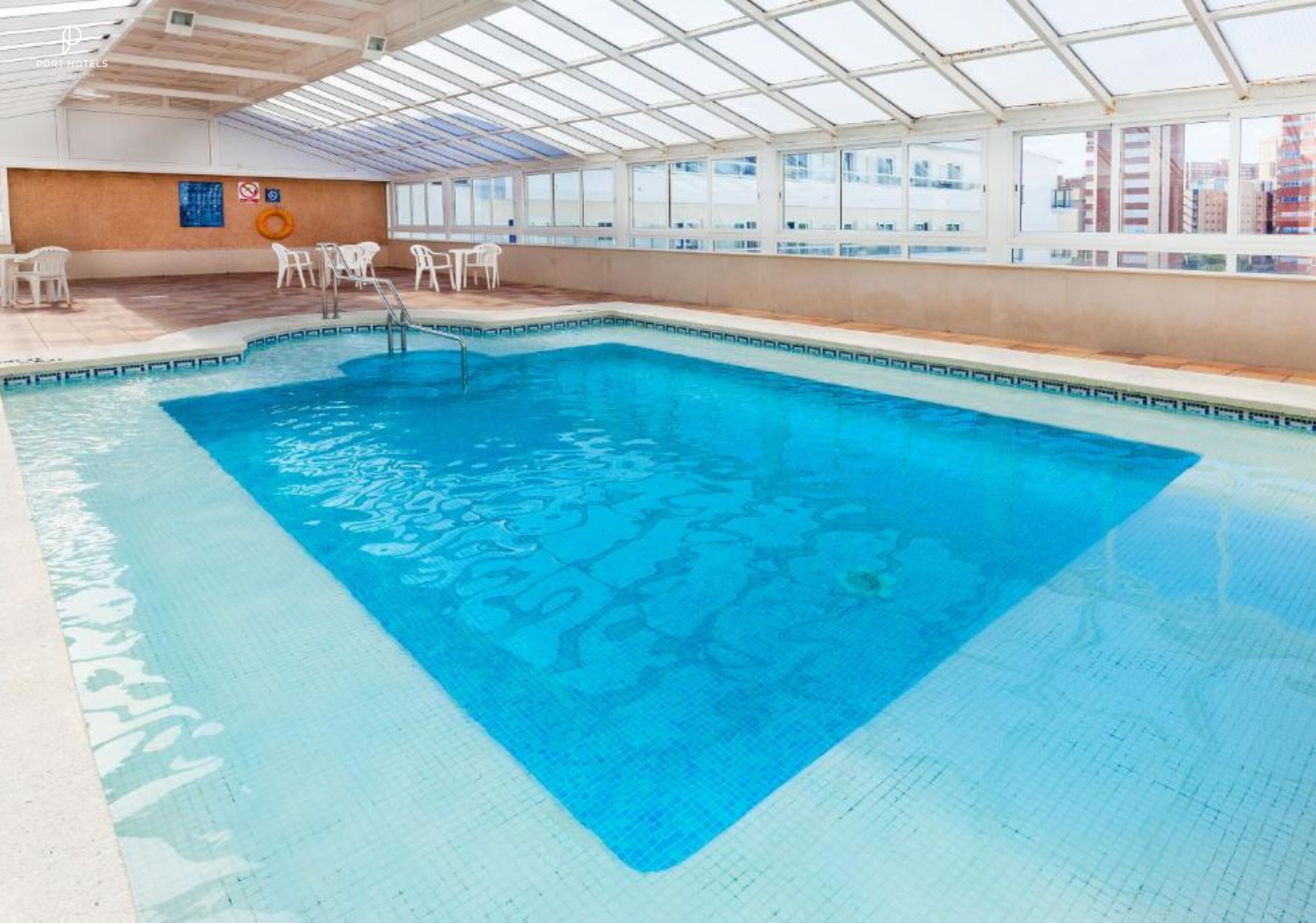An indoor swimming pool with clear blue water, surrounded by white chairs and tables, and large windows offering a view of the city.