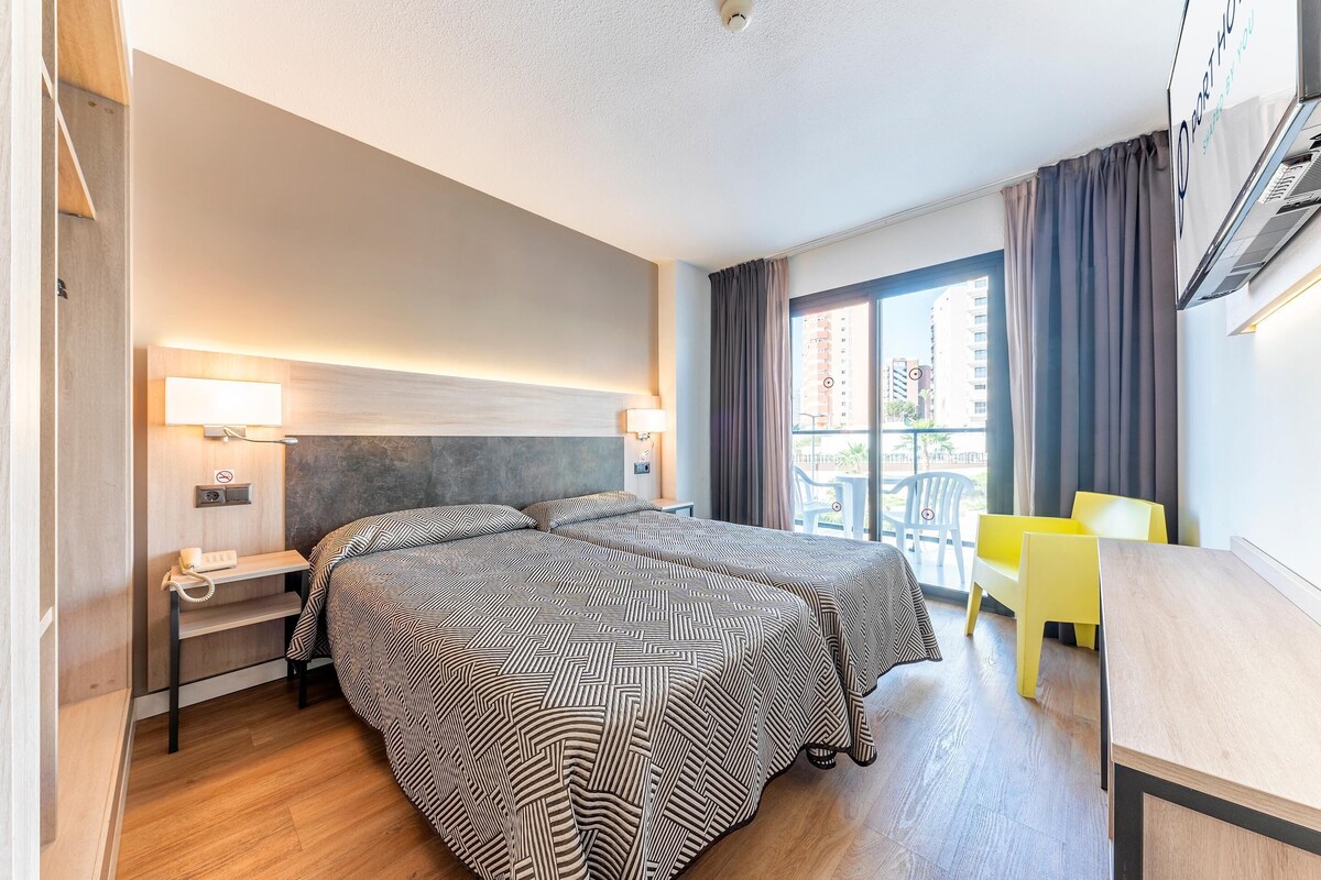 A hotel room with a bed, bedside table, lamp, and a view of the outside balcony.