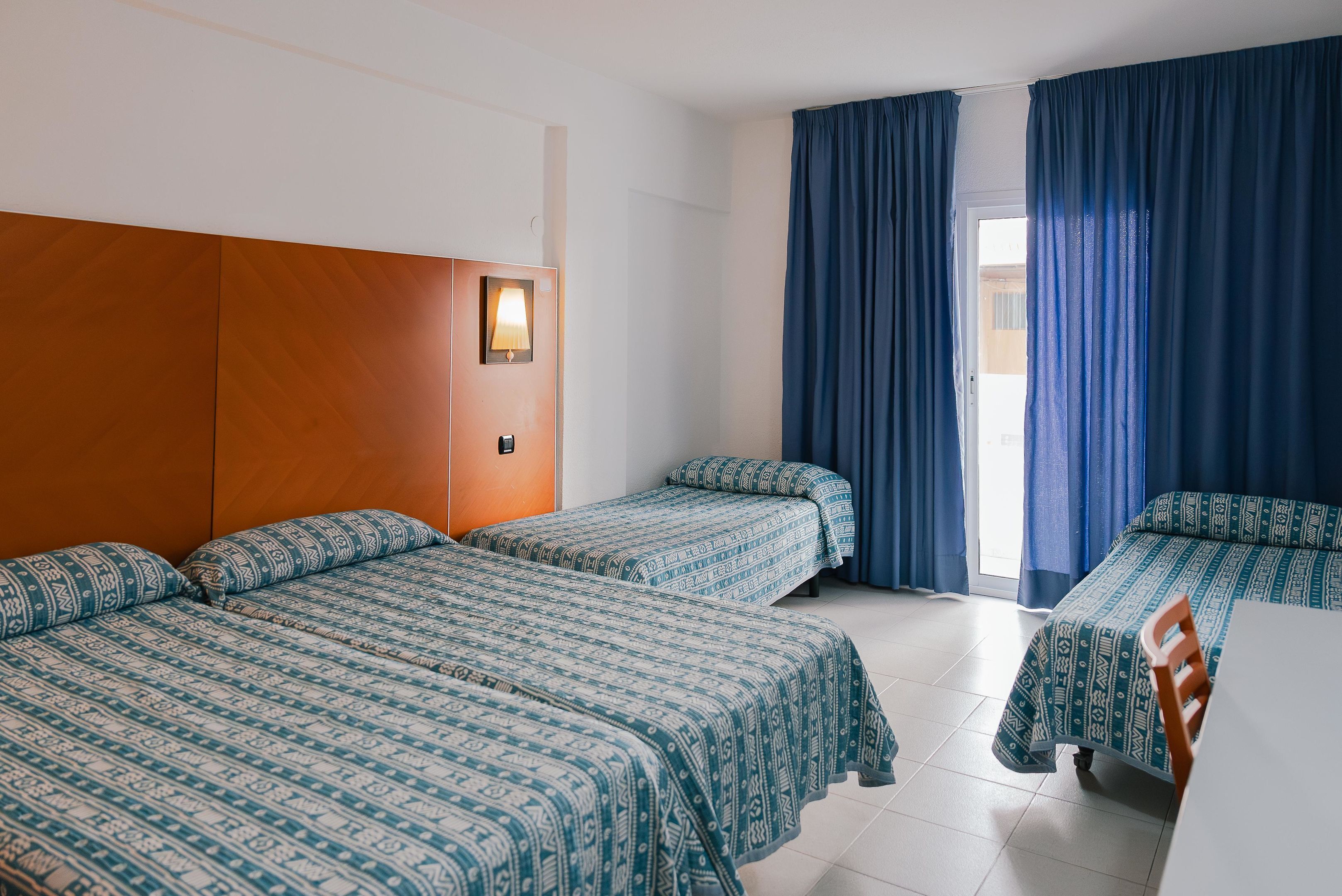 A hotel room with two single beds, a wooden headboard, a bedside table, and a window with blue curtains.
