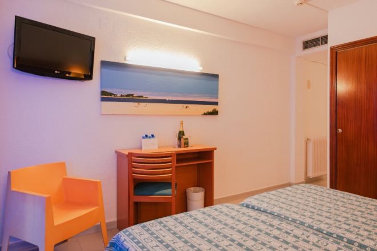 A hotel room with a bed, a TV mounted on the wall, a wooden desk with a chair, and a painting of a beach scene.