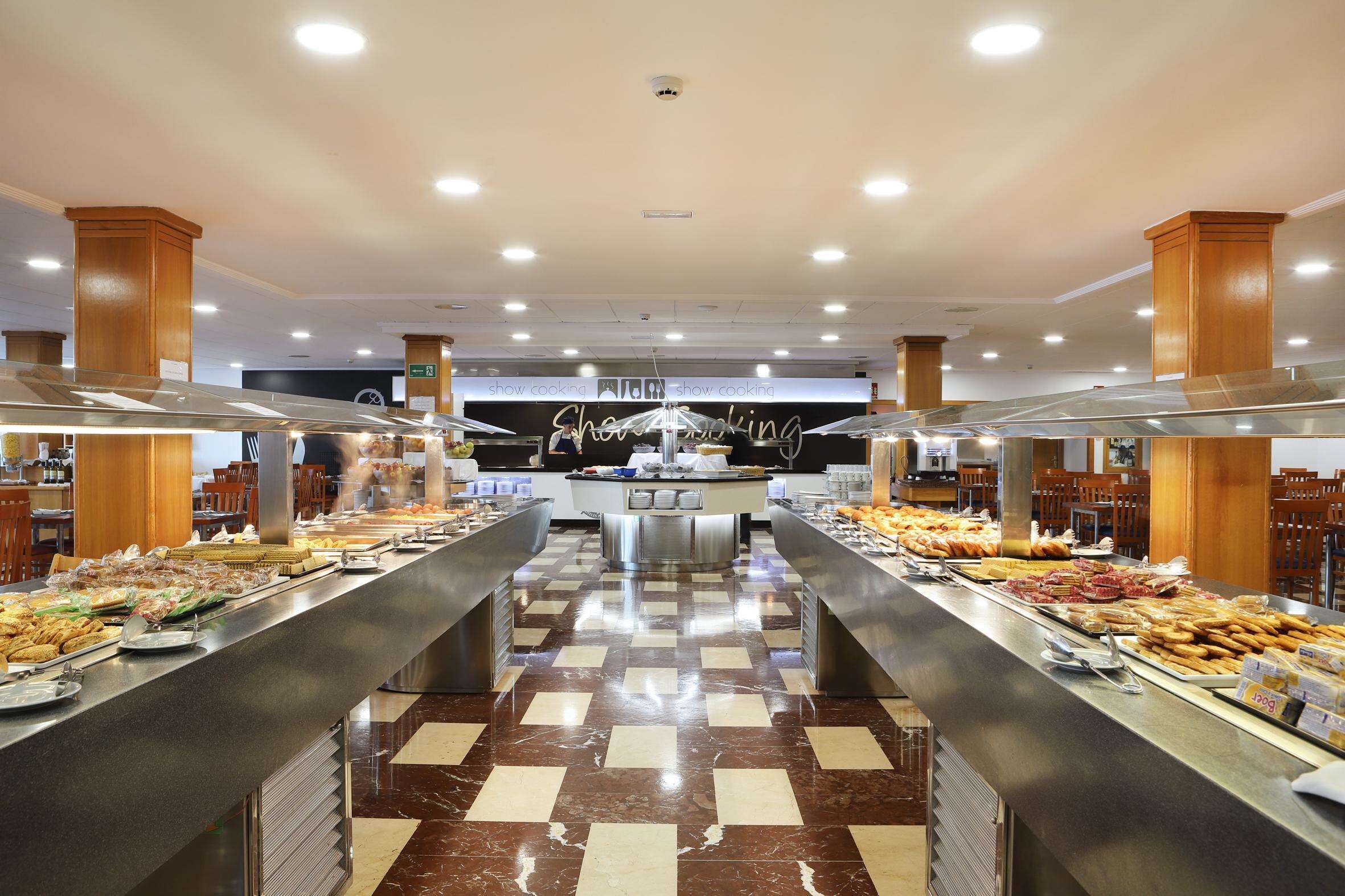 Un espace buffet avec plusieurs stands de nourriture, un îlot central sur lequel est affiché un panneau indiquant « démonstration culinaire », et un comptoir d’enregistrement en arrière-plan.