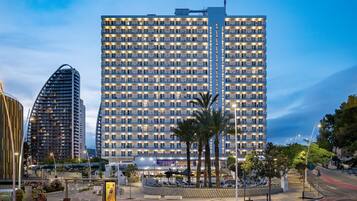 A high-rise hotel with multiple balconies, a central vertical feature, and a well-lit urban street with palm trees and benches.