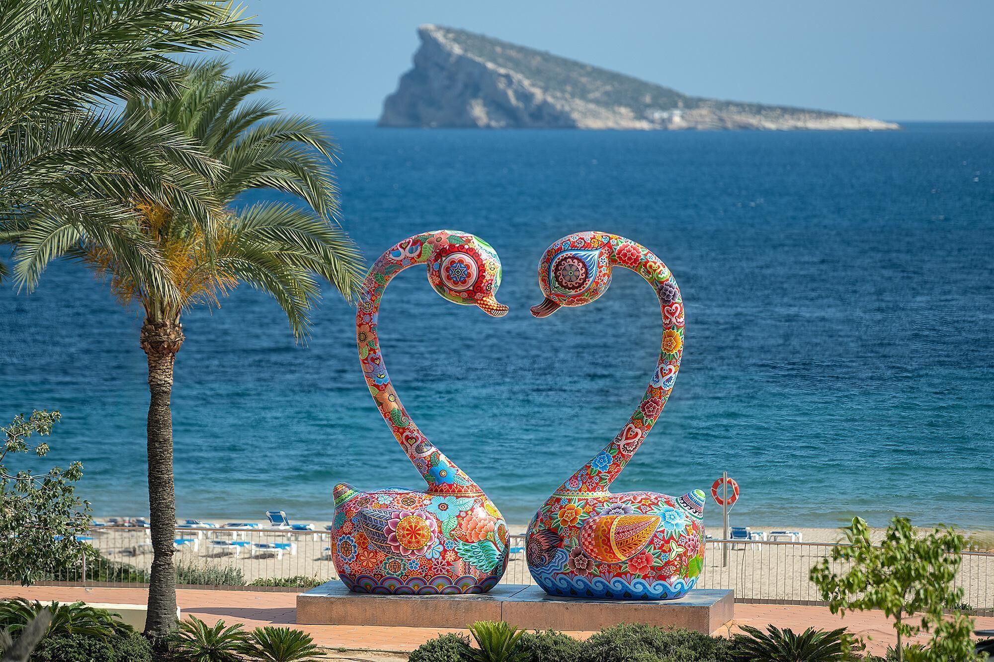 Eine dekorative Skulptur von zwei Schwänen mit herzförmigen Hälsen vor einer Strand- und Ozeanlandschaft.