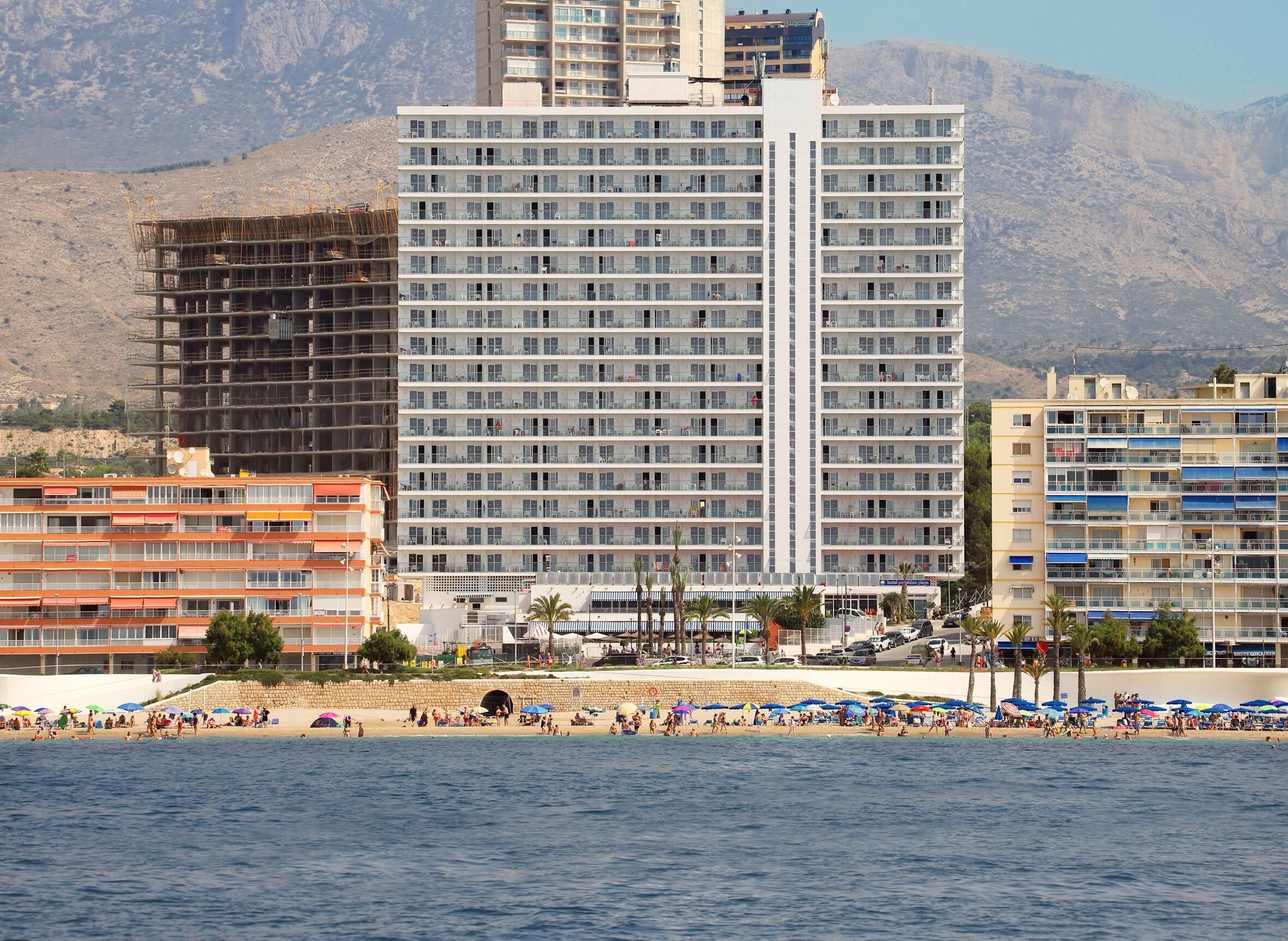A coastal hotel with multiple floors, a beach in the foreground, and mountains in the background.