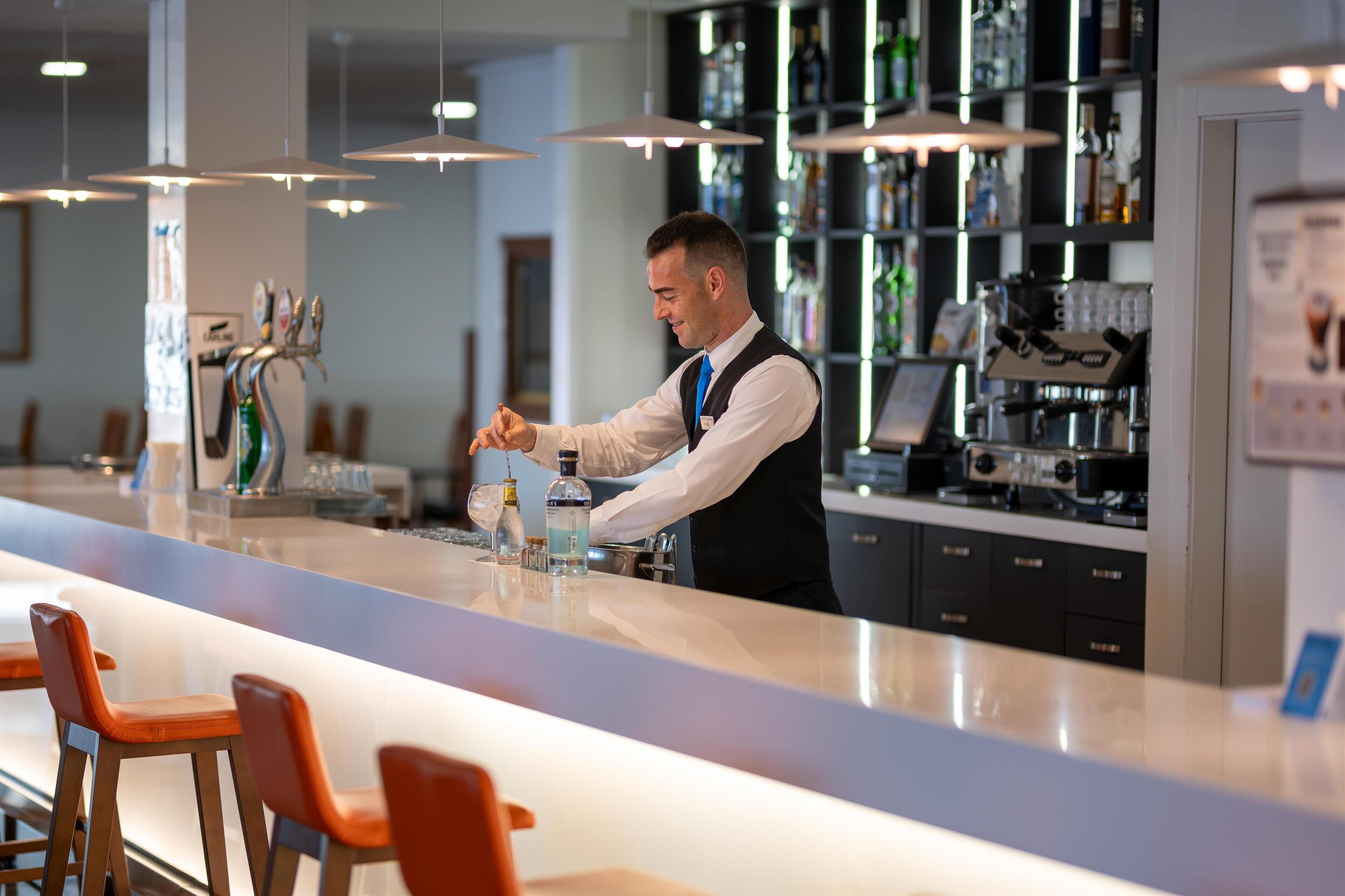 A modern bar area with a bartender preparing drinks, bar stools, and a well-stocked shelf with various bottles.