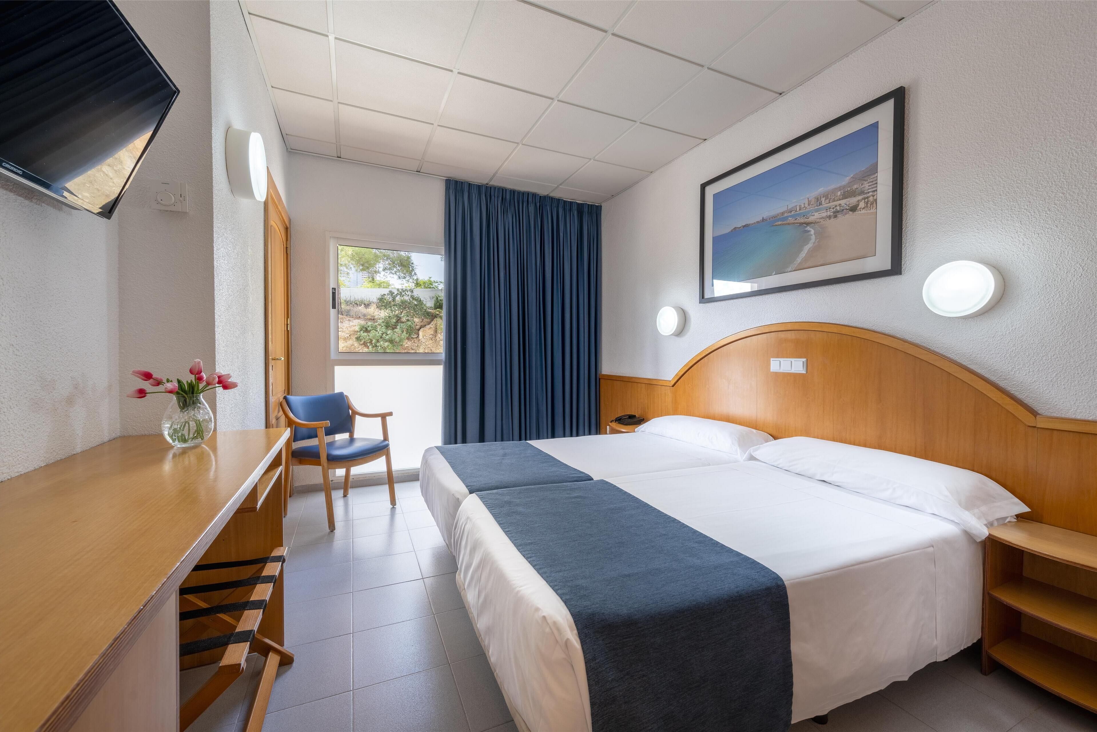 A hotel room with a bed, a desk, a chair, a TV, a painting, and a window with curtains.