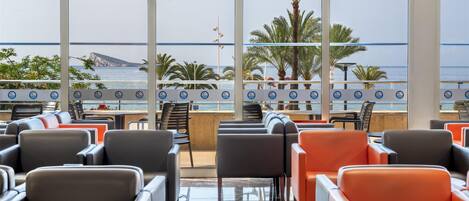 Vestíbulo de hotel con amplias ventanas que dan a una playa, con filas de sillas y una mesa central.