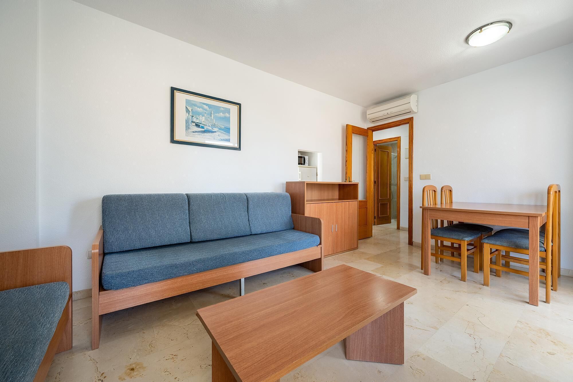 A living room with a sofa, a wooden coffee table, a dining table with chairs, a framed picture on the wall, and an air conditioning unit.