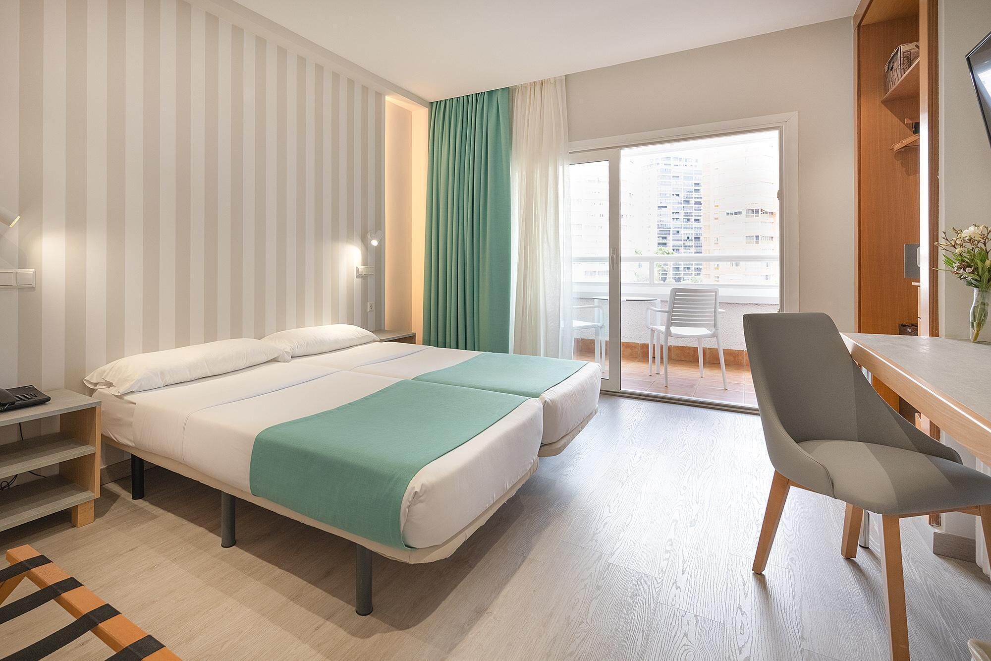 Een hotelkamer met een bed, een bureau, een stoel en uitzicht op de stad door een groot raam.