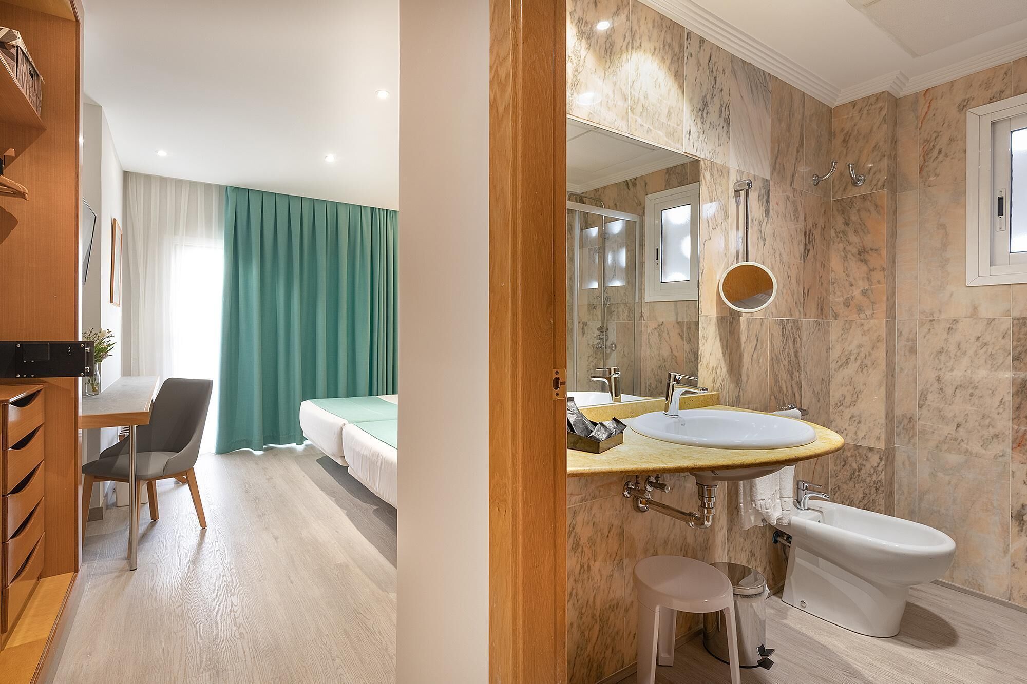 A hotel room with a wooden desk, a bed with green curtains, a bathroom with a marble wall, a sink, and a bidet.