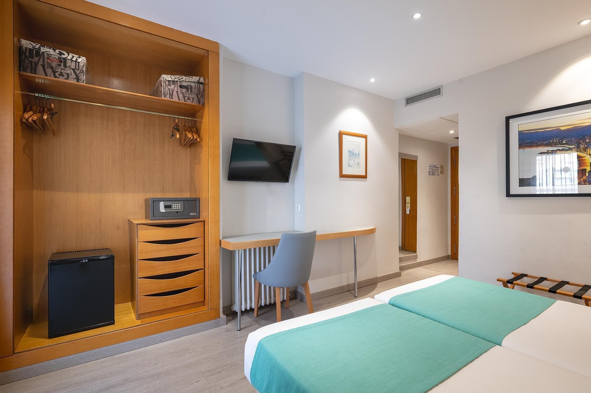 A hotel room with a bed, a desk with a chair, a wardrobe, a TV, and a framed picture on the wall.