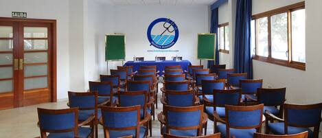 Un salón de conferencias con filas de sillas de madera y una mesa, una pizarra y un logotipo en la pared.