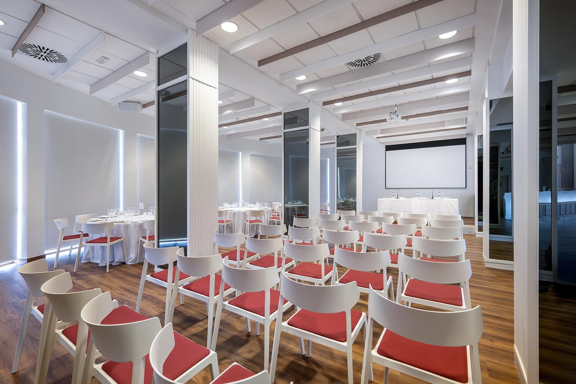 Une salle de conférence avec des rangées de chaises blanches dotées de coussins rouges, un grand écran blanc et des tables disposées pour une réunion.