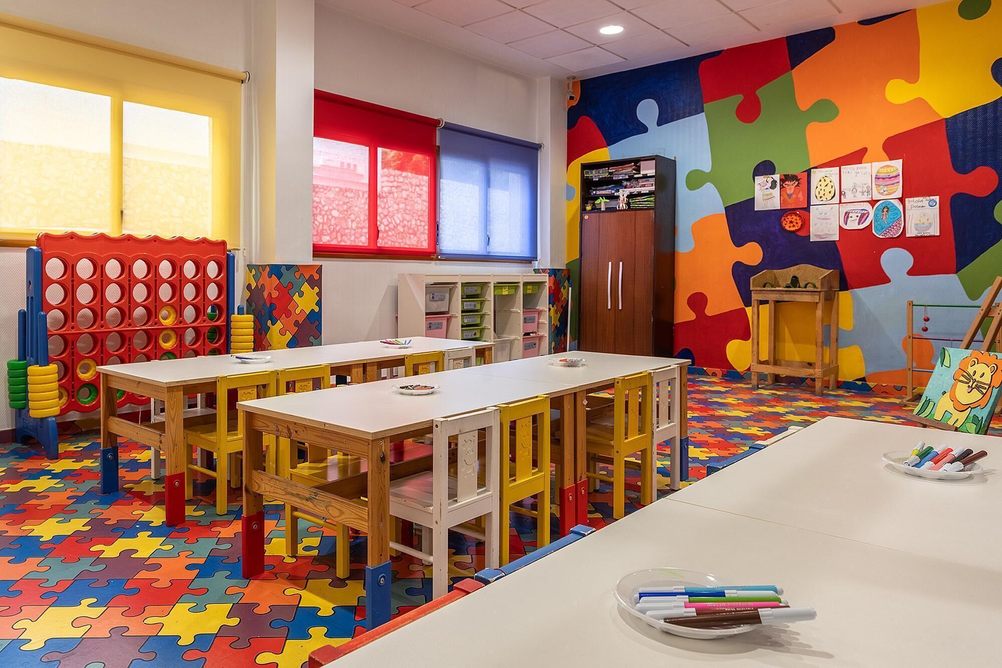 Une salle de classe dotée d’un sol coloré en forme de puzzle, de tables et de chaises en bois, d’une bibliothèque et d’une fenêtre avec des stores rouges.