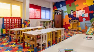 Une salle de classe dotée d’un sol coloré en forme de puzzle, de tables et de chaises en bois, d’une bibliothèque et d’une fenêtre avec des stores rouges.