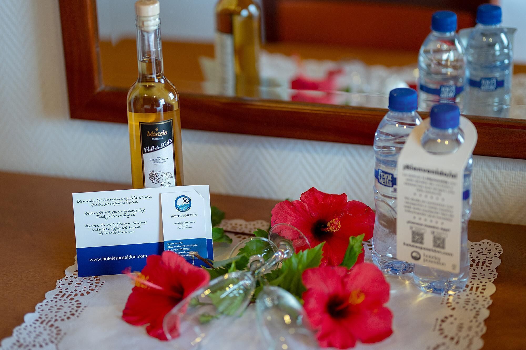 A hotel room with a bottle of wine, water bottles, a welcome card, and red flowers on a table.