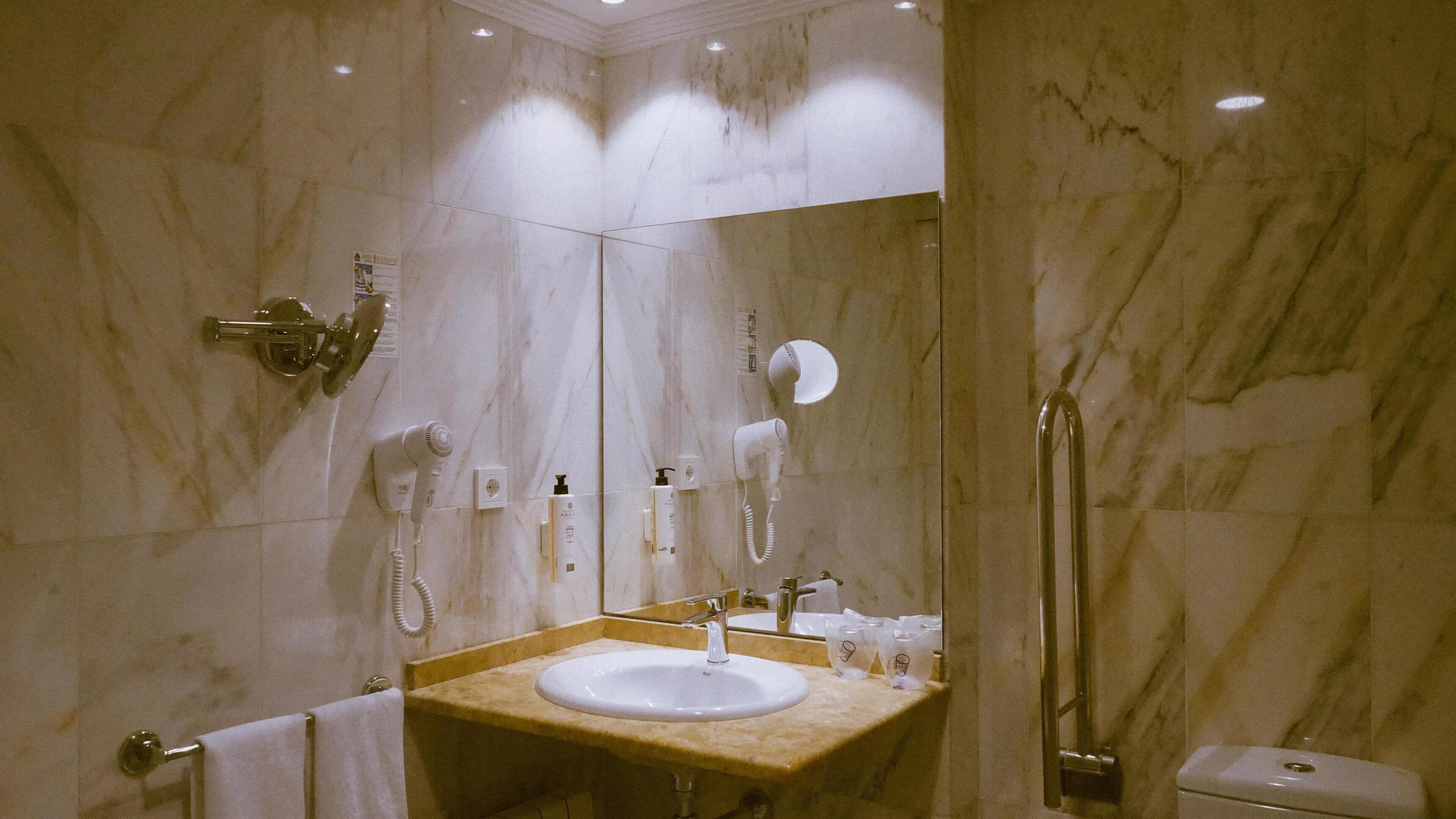 A bathroom with a marble wall, a sink, a mirror, a hairdryer, and a towel rack.