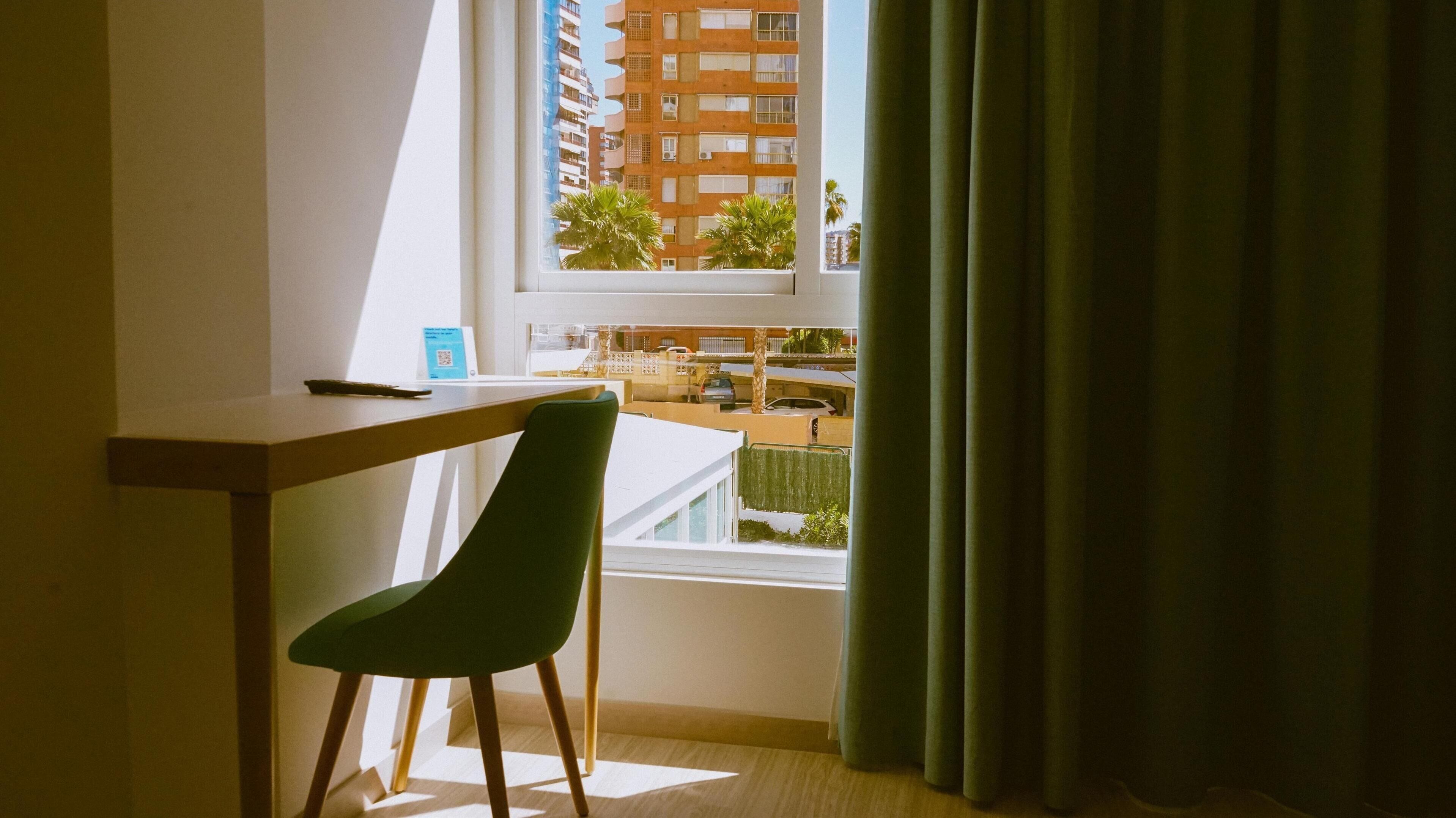 A room with a desk, a chair, a window with curtains, and a view of a building and palm trees outside.