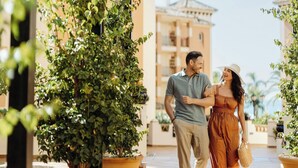 Una pareja caminando bajo una pérgola de madera con plantas en macetas y un edificio al fondo.