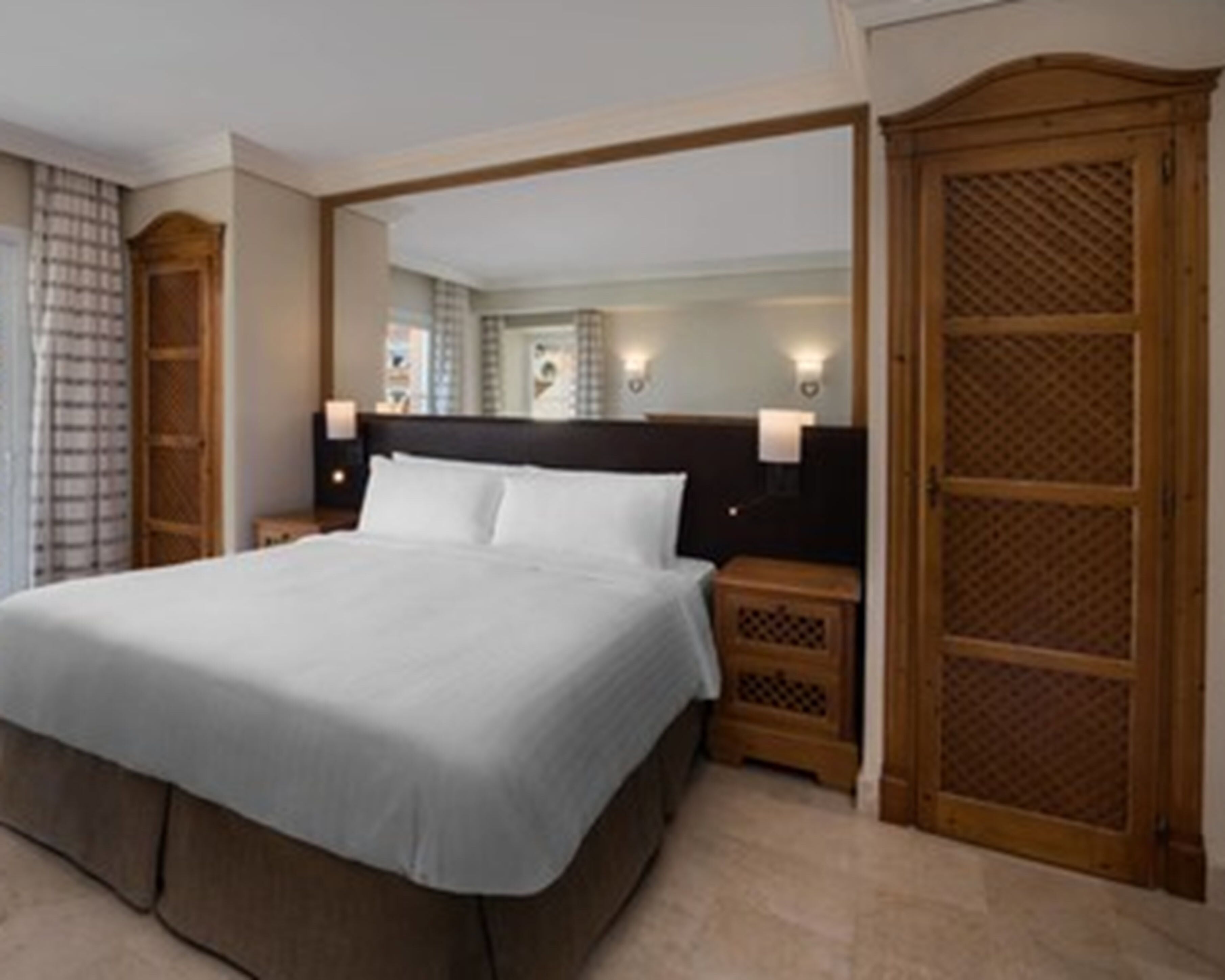 A hotel room with a large bed, wooden headboard, two bedside tables with lamps, a mirror, and a wooden wardrobe.