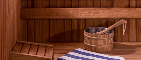 Una sauna de madera con un banco, un cubo de metal y una cuchara de madera, y una toalla a rayas sobre el banco.