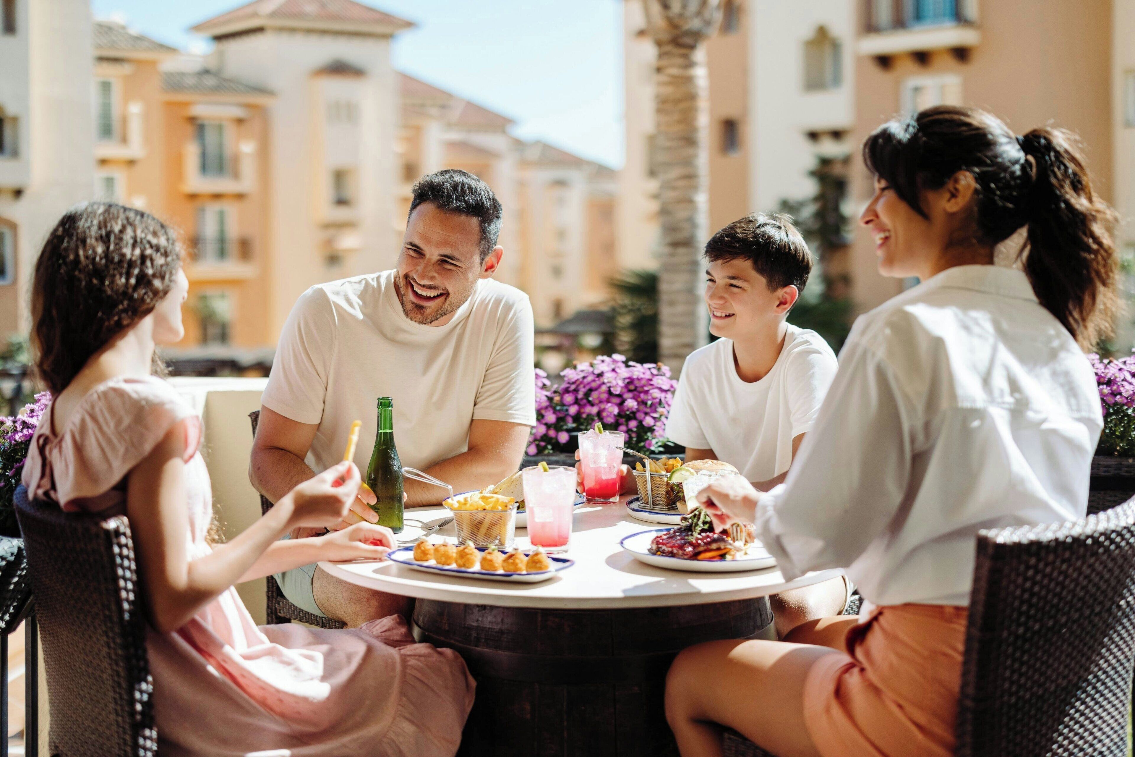 Um grupo de pessoas jantando ao ar livre em uma mesa redonda, com comida e bebidas, cercado por edifícios e plantas.