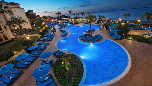Zona de piscina en un resort con sillones de descanso y sombrillas, rodeada de palmeras y vista al océano.