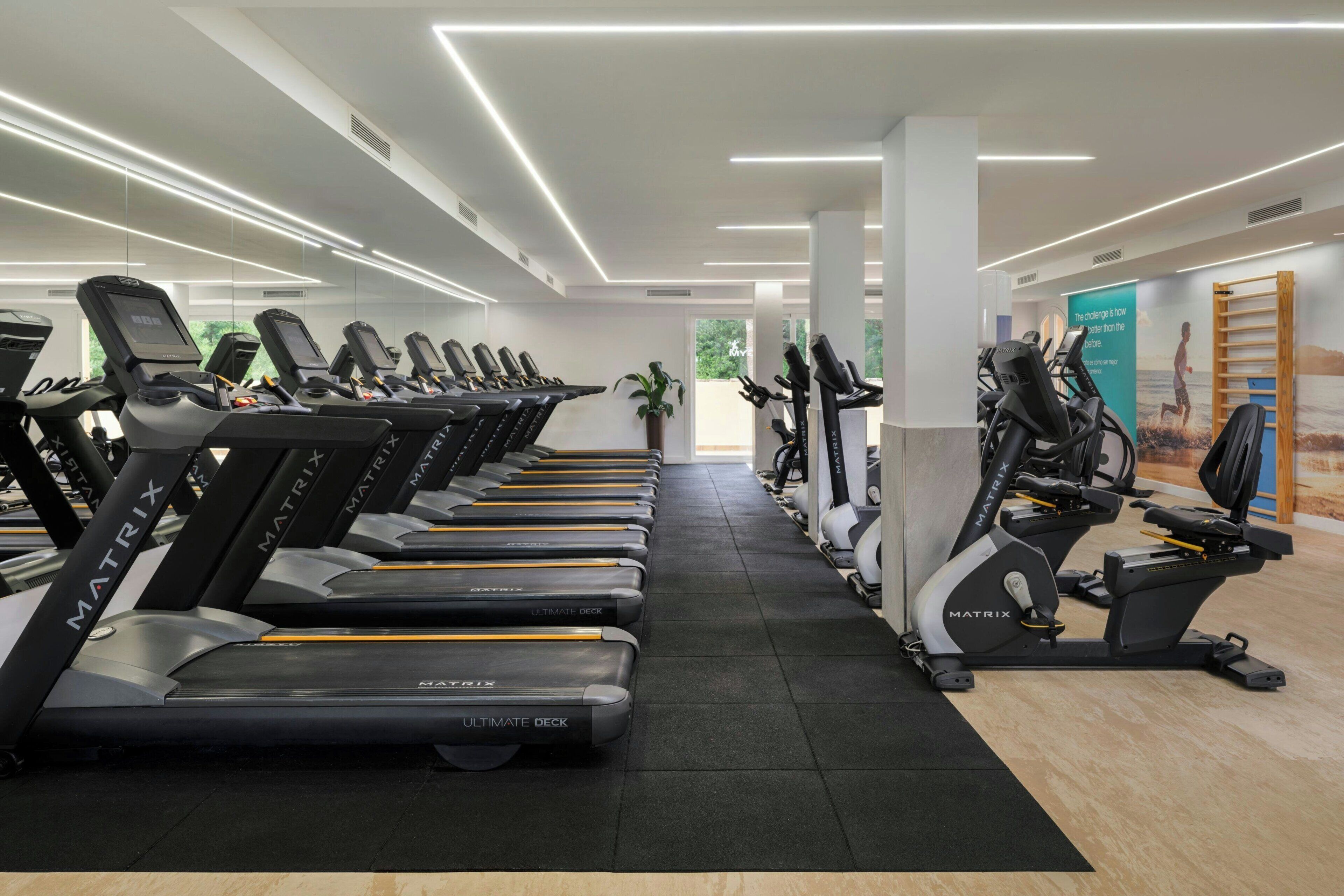Une salle de sport équipée de plusieurs tapis de course et de plusieurs appareils elliptiques, avec une décoration aux couleurs noir et blanc et une vue sur l’extérieur grâce à de grandes fenêtres.