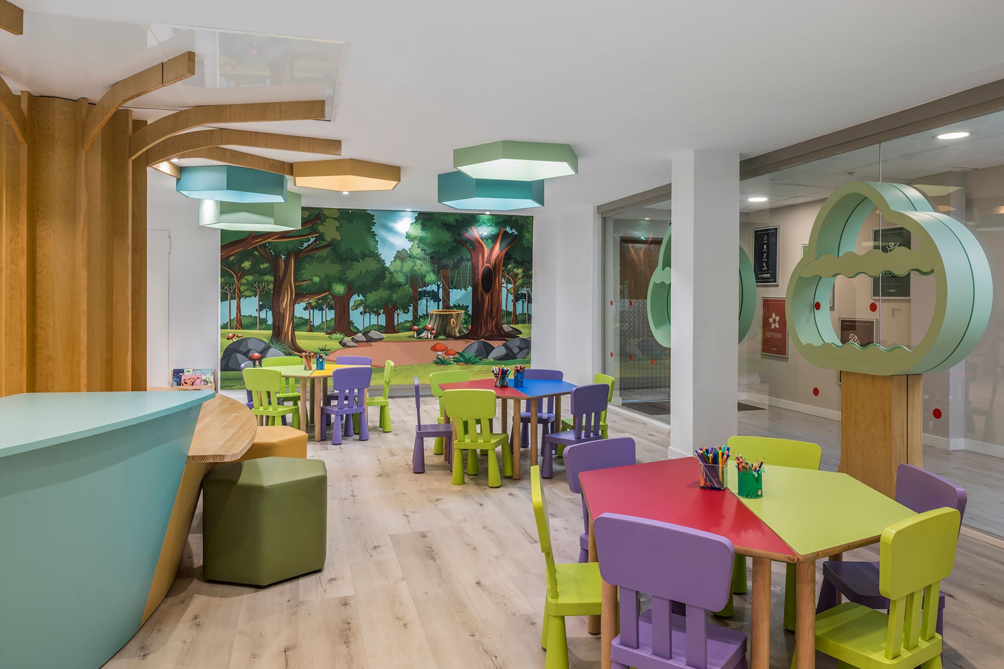 Un espace de jeux pour enfants avec des chaises et des tables colorées, une fresque représentant une forêt et un comptoir d’accueil en bois.