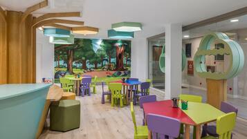 Un espace de jeux pour enfants avec des chaises et des tables colorées, une fresque représentant une forêt et un comptoir d’accueil en bois.