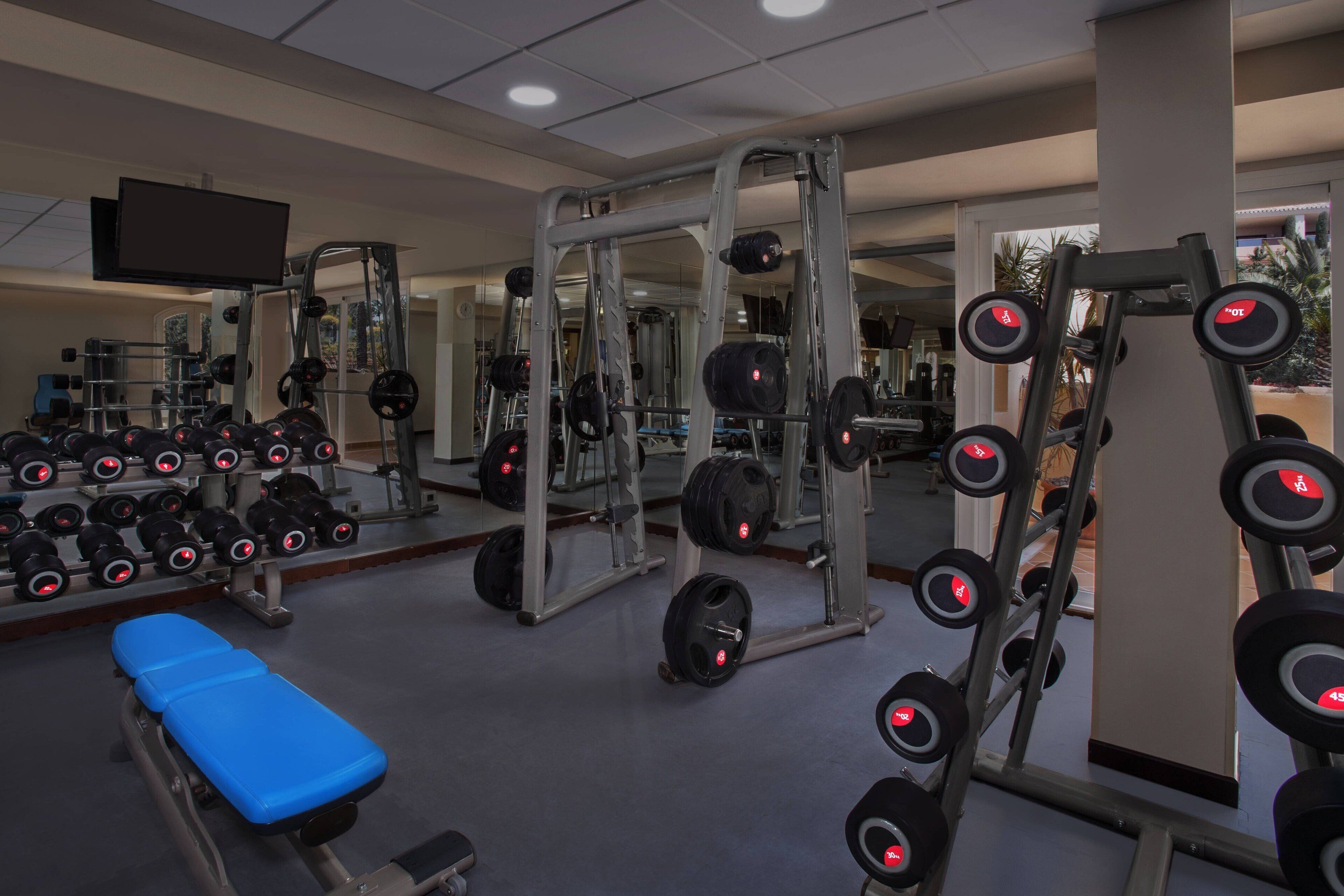 A gym with weightlifting equipment, benches, and a television screen.