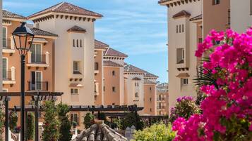 Un pati amb edificis amb teulades de terracota i balcons, envoltat de verdura exuberant i flors rosades vibrants.