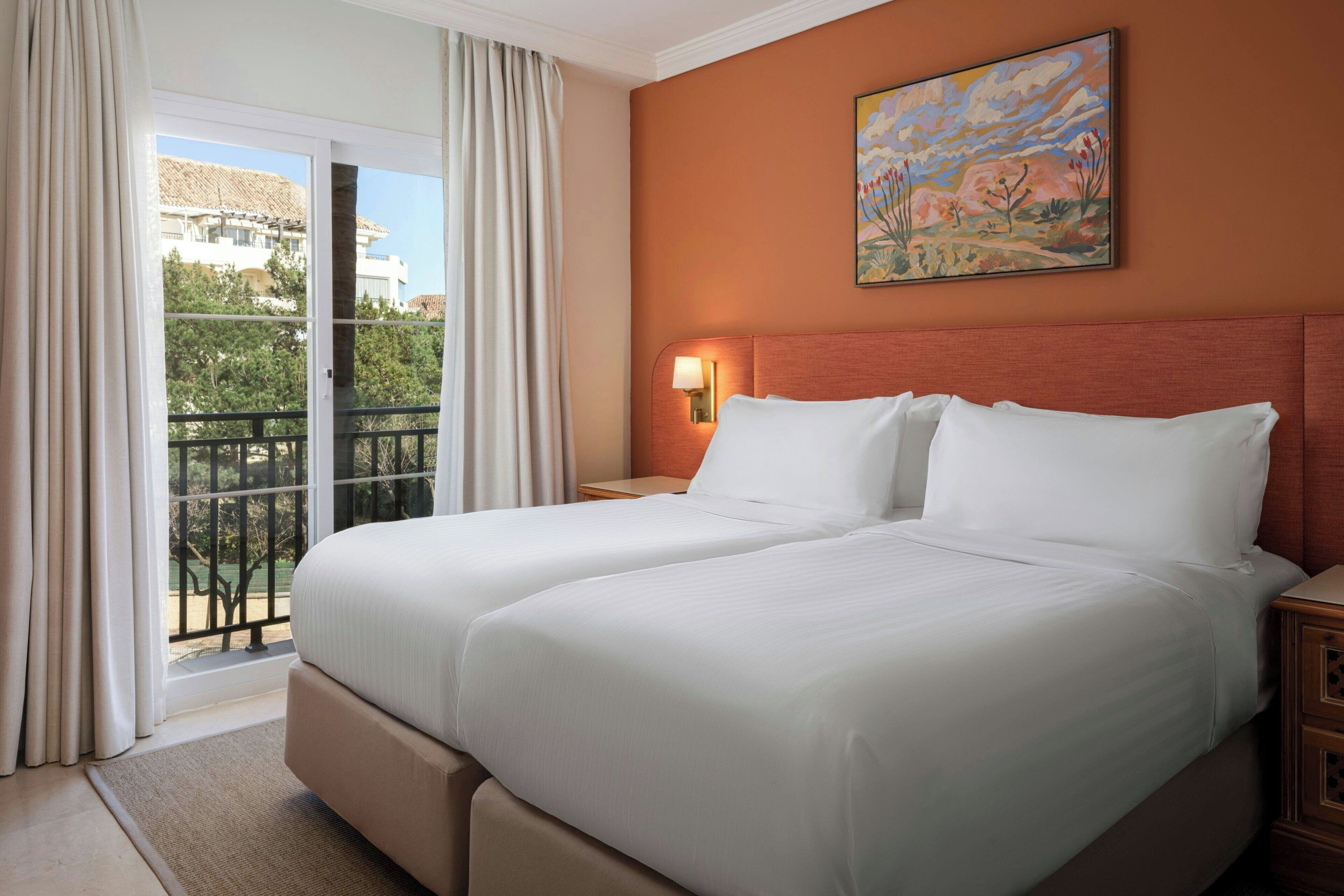 Habitación de hotel con dos camas, un balcón, un cuadro en la pared y una mesita de noche con lámpara.