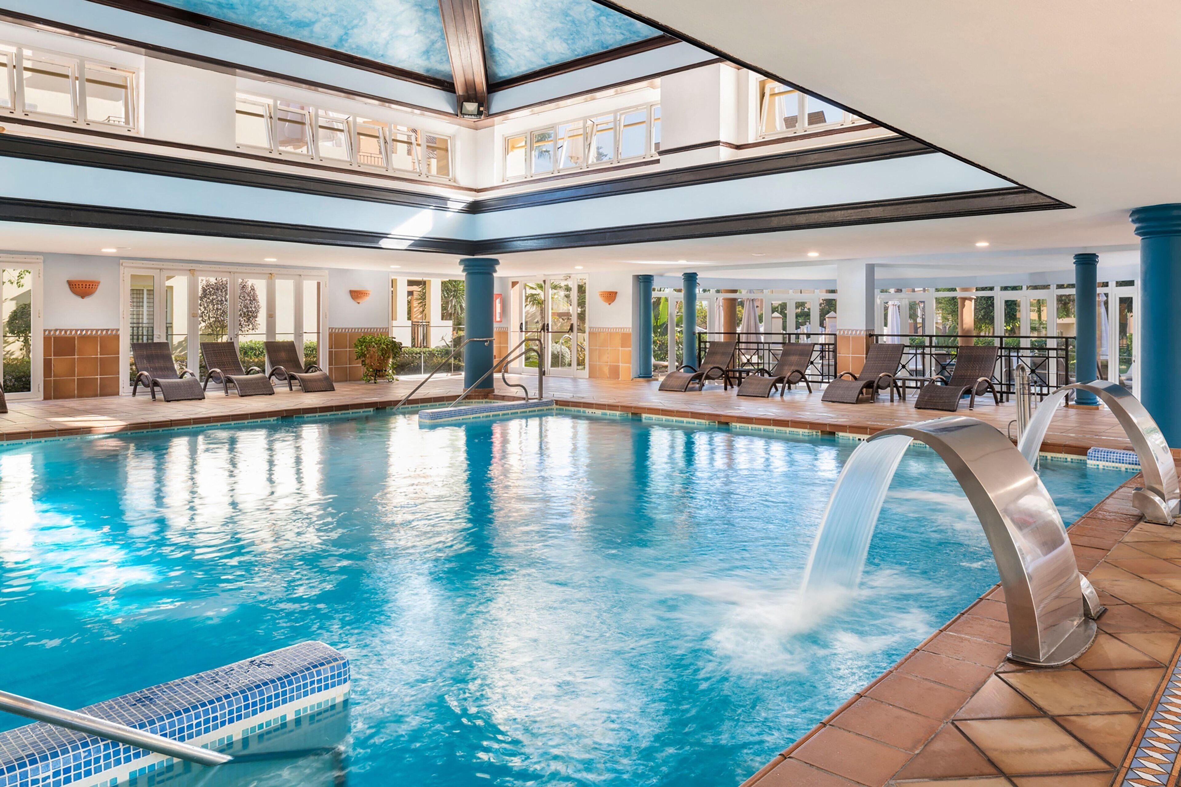 Una piscina interior con caída de agua, varias zonas de descanso y vistas al exterior a través de grandes ventanales.