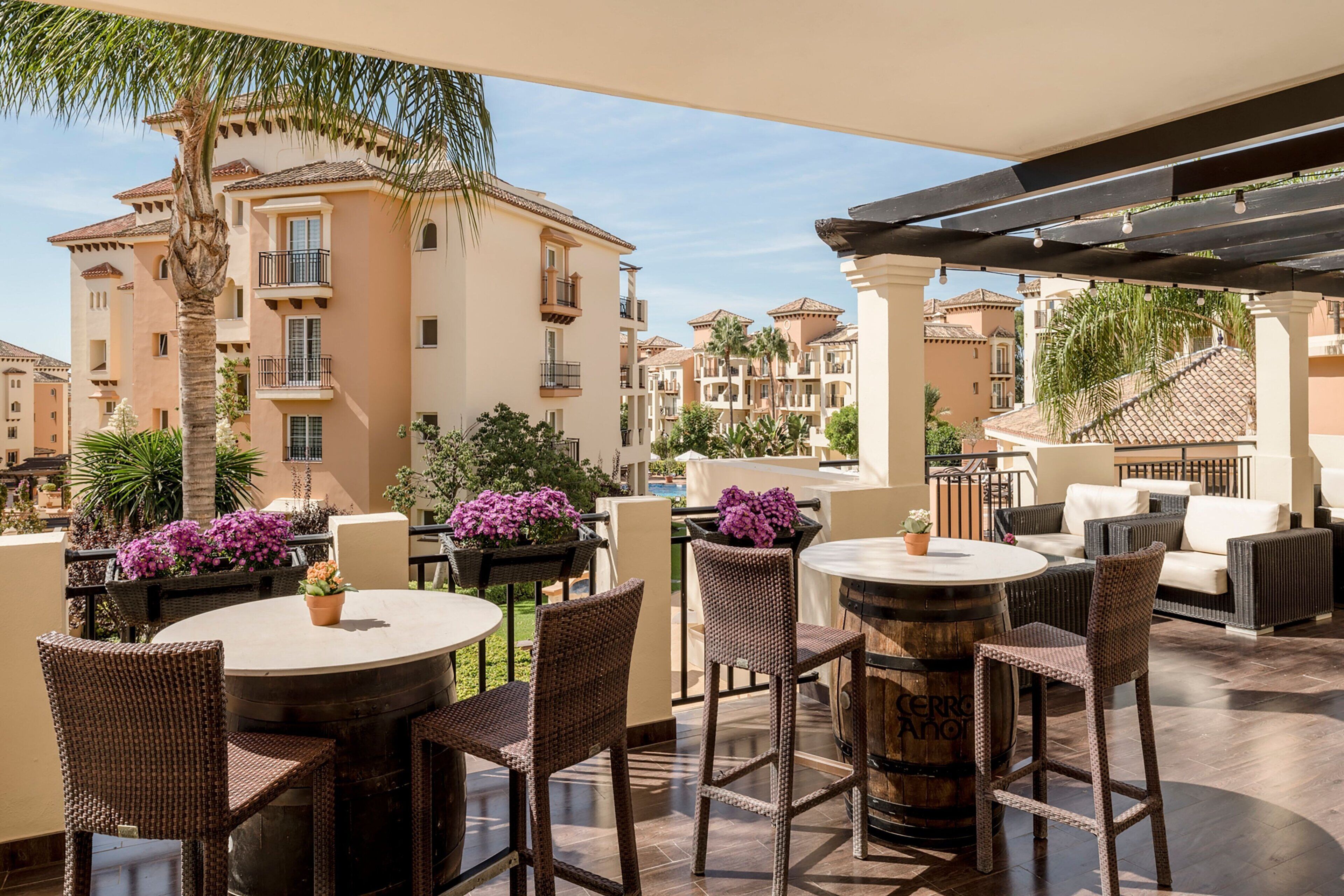 Un espace extérieur aménagé en terrasse, avec des tables rondes et des chaises en osier, entouré de plantes en pot et offrant une vue sur des immeubles à plusieurs étages.