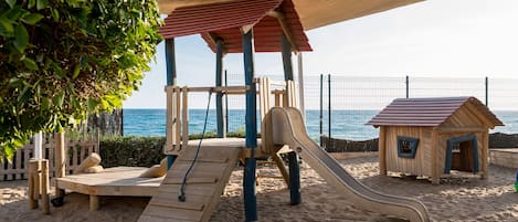 A beach playground with a slide and climbing structures, a wooden playhouse, and a covered seating area.