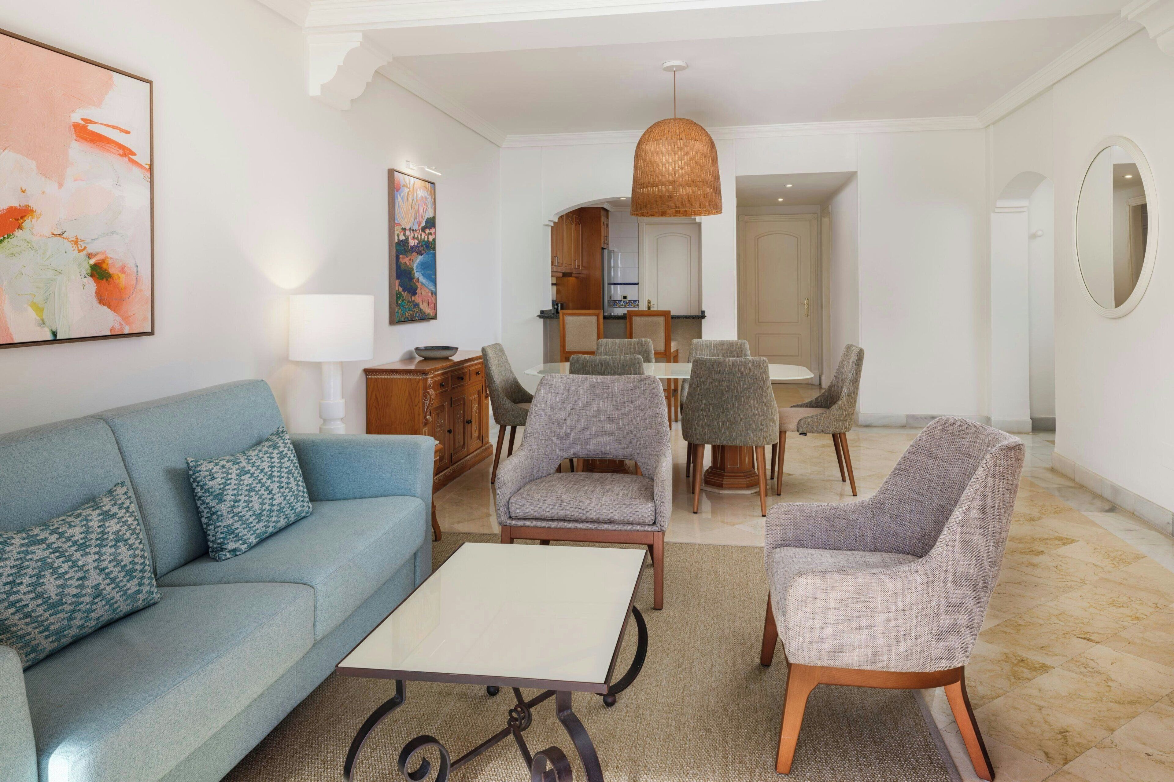 A living room with a sofa, armchairs, a coffee table, a dining area with chairs, a kitchen in the background, and a large painting on the wall.